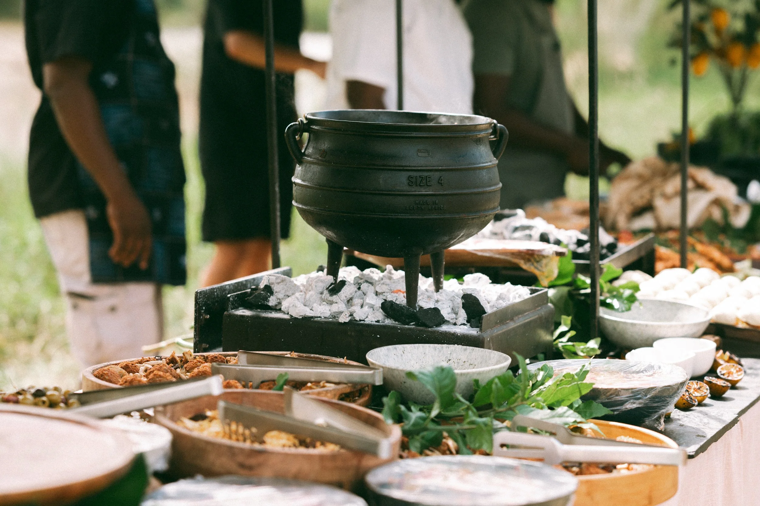 Outdoor buffet with various dishes on tables and a black pot over coals