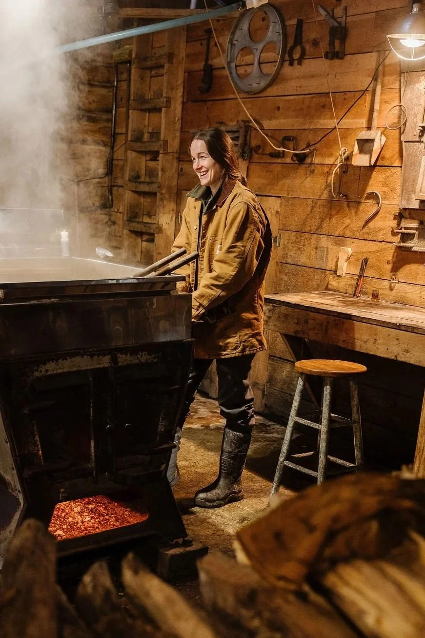 Woman in a brown jacket and rubber boots standing in a rustic wooden workshop near a steaming stove, surrounded by woodworking tools and stacked firewood.