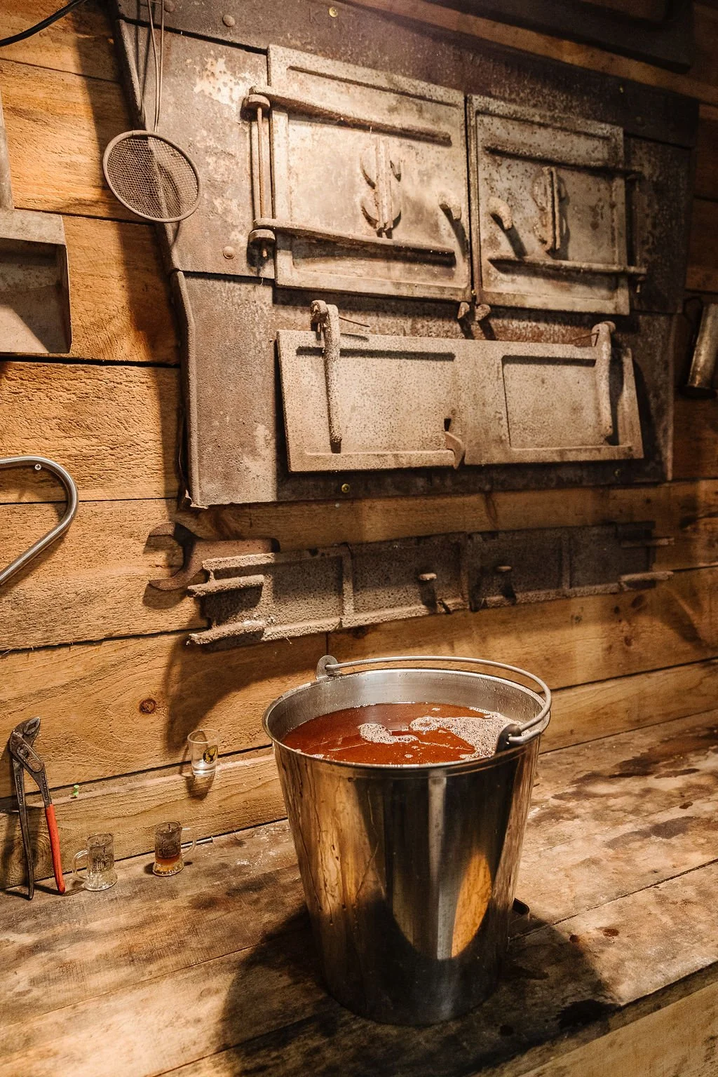 A metal bucket filled with a dark liquid on a wooden table, with an antique wood stove in the background and small shot glasses on the table.