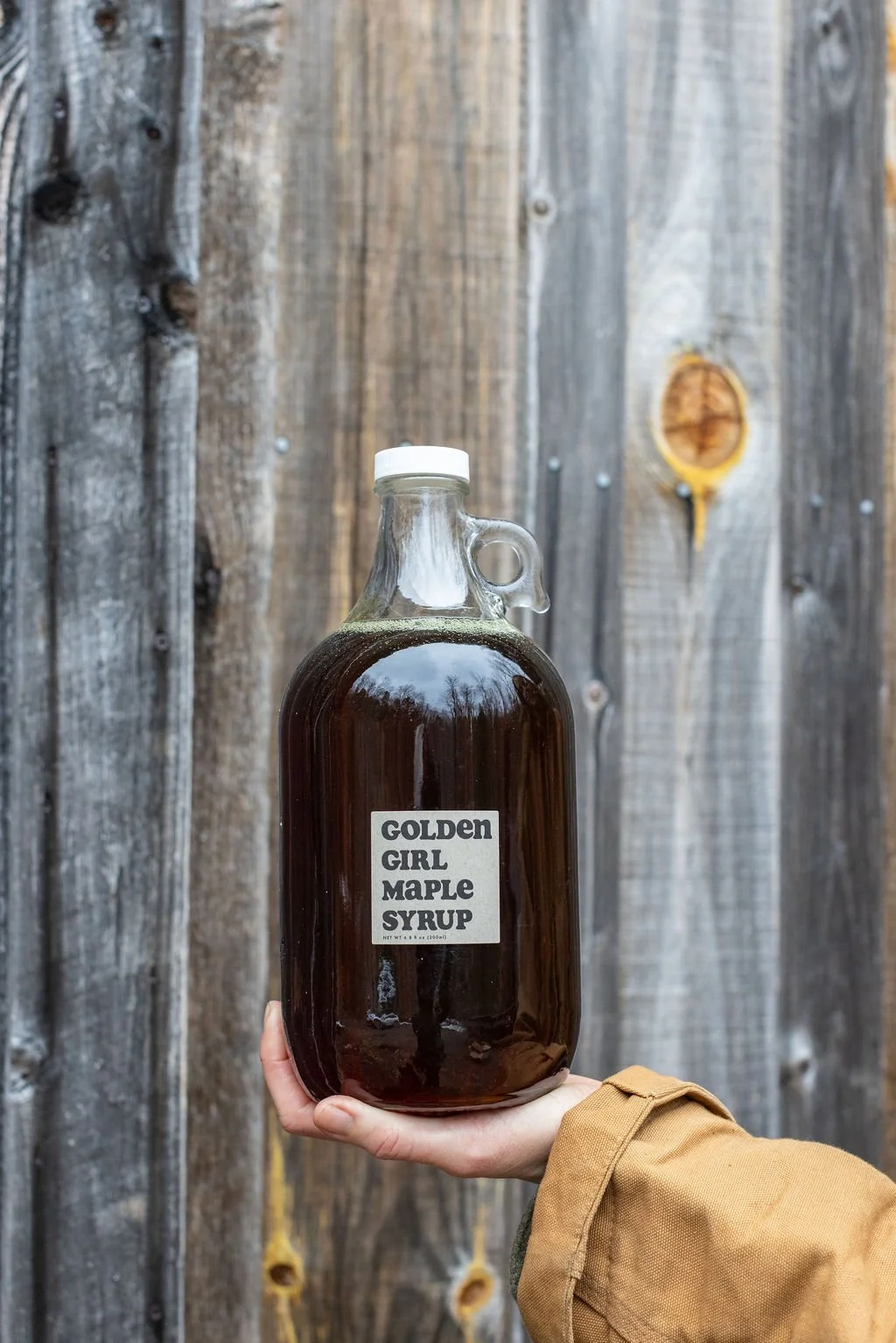 A person holding a glass jug of dark maple syrup labeled 'Golden Girl Maple Syrup' in front of a wooden barn.
