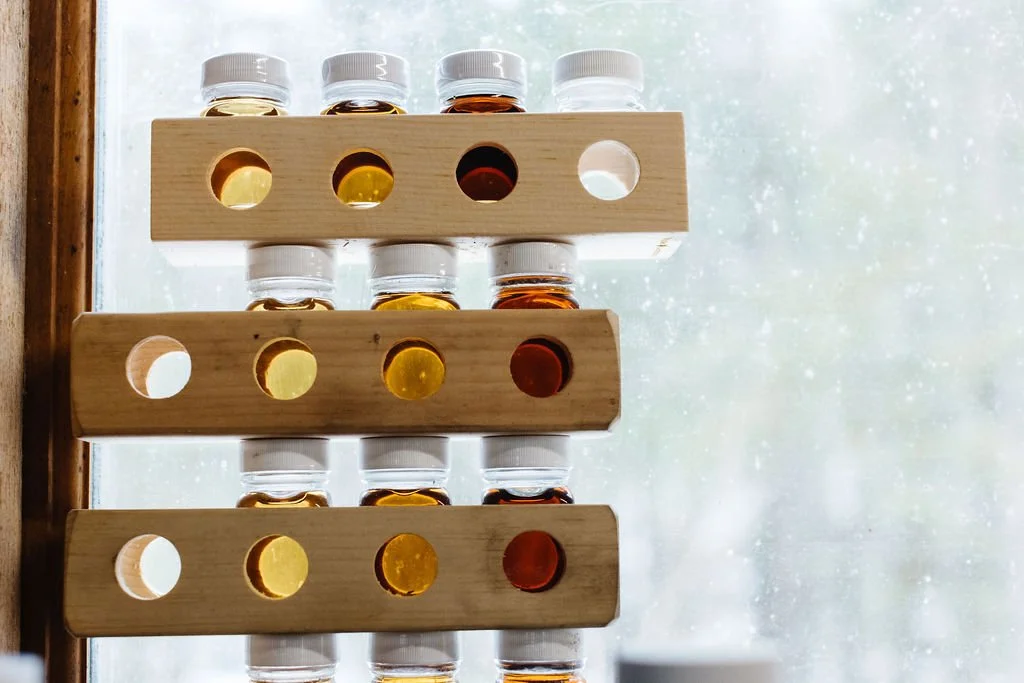 Three wooden spice racks holding small glass jars with white caps, each containing different colored liquids, placed next to a window with a rainy scene outside.