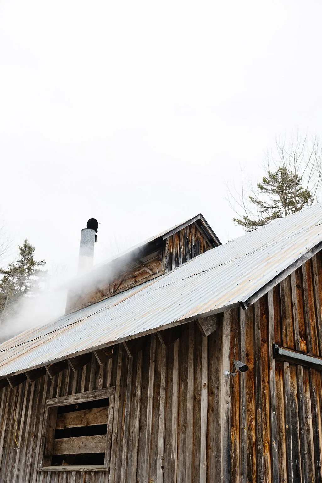 A rustic wooden barn with a sloped metal roof, smoke coming out of a chimney, and leafless trees in the background under a cloudy sky.