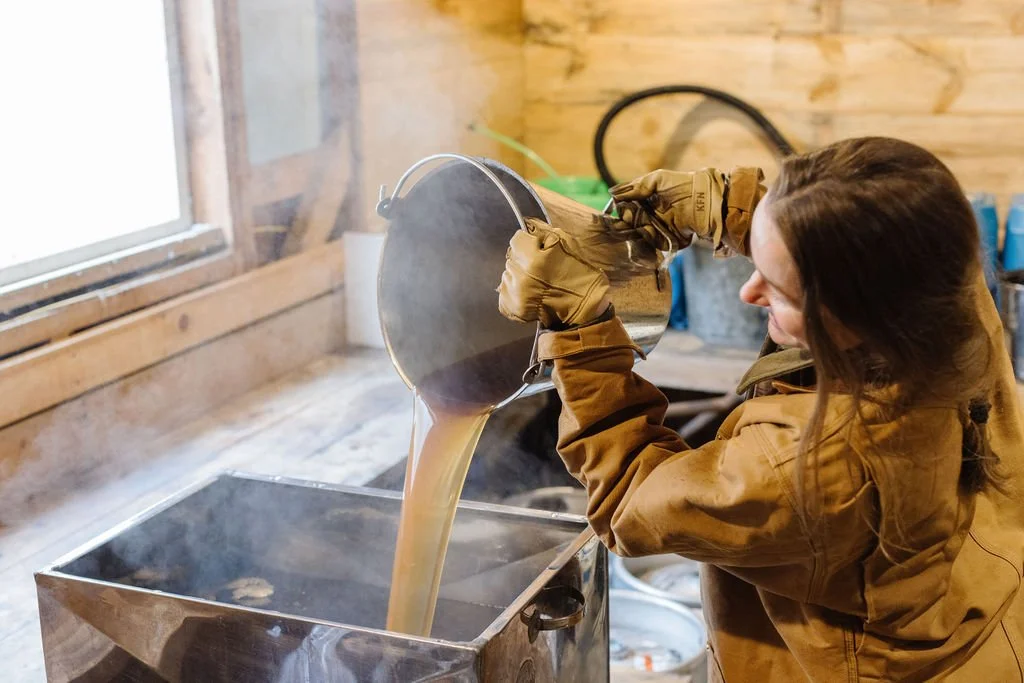 Woman pouring hot liquid from a metal container into a rectangular metal vat, with steam rising, inside a wooden cabin or workshop.