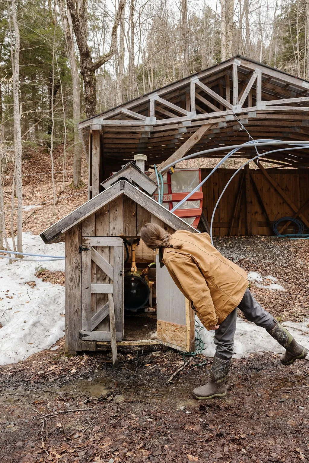 A person in outdoor clothing inspecting an outdoor structure with pipes and equipment, surrounded by trees and snow on the ground.