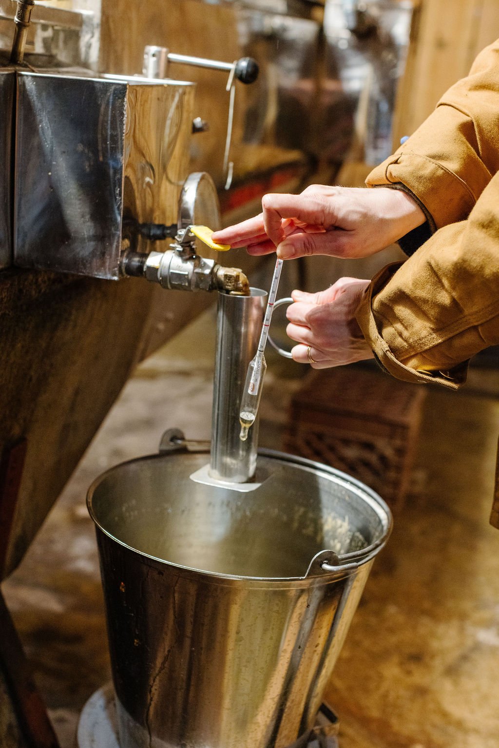 A person in a Carhartt jacket draws off hot maple syrup into a stainless steel bucket out of an evaporator holding a hydrometer.