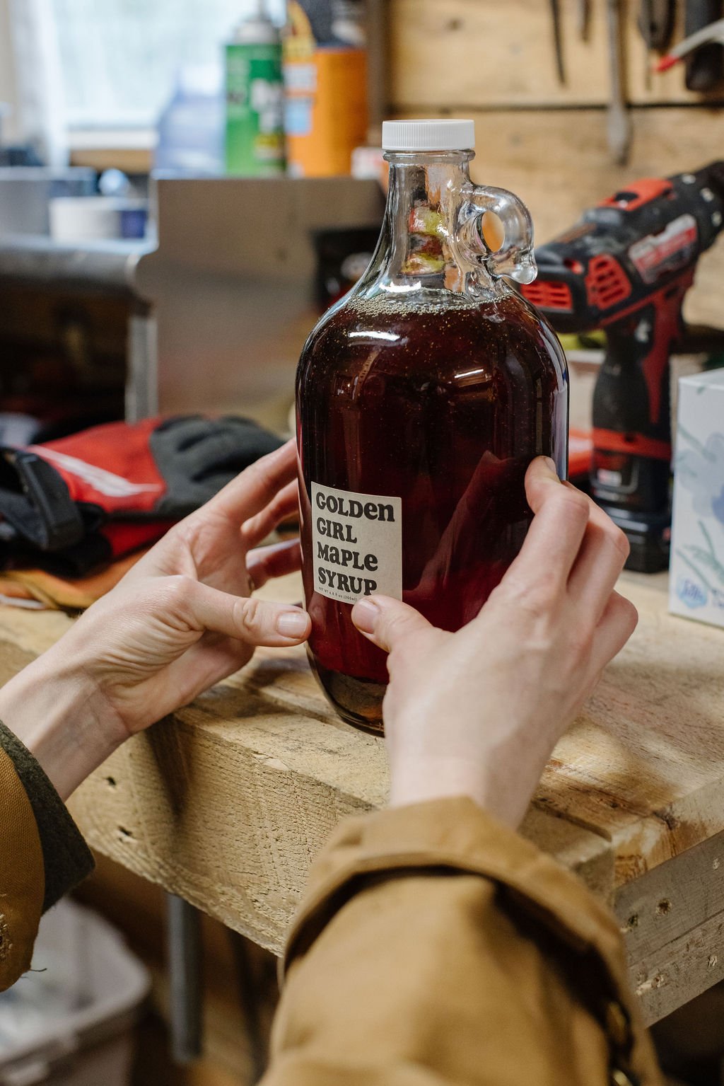 Person holding a glass bottle labeled 'GOLDEN GIRL MAPLE SYRUP' filled with dark syrup, in a workshop or garage setting.