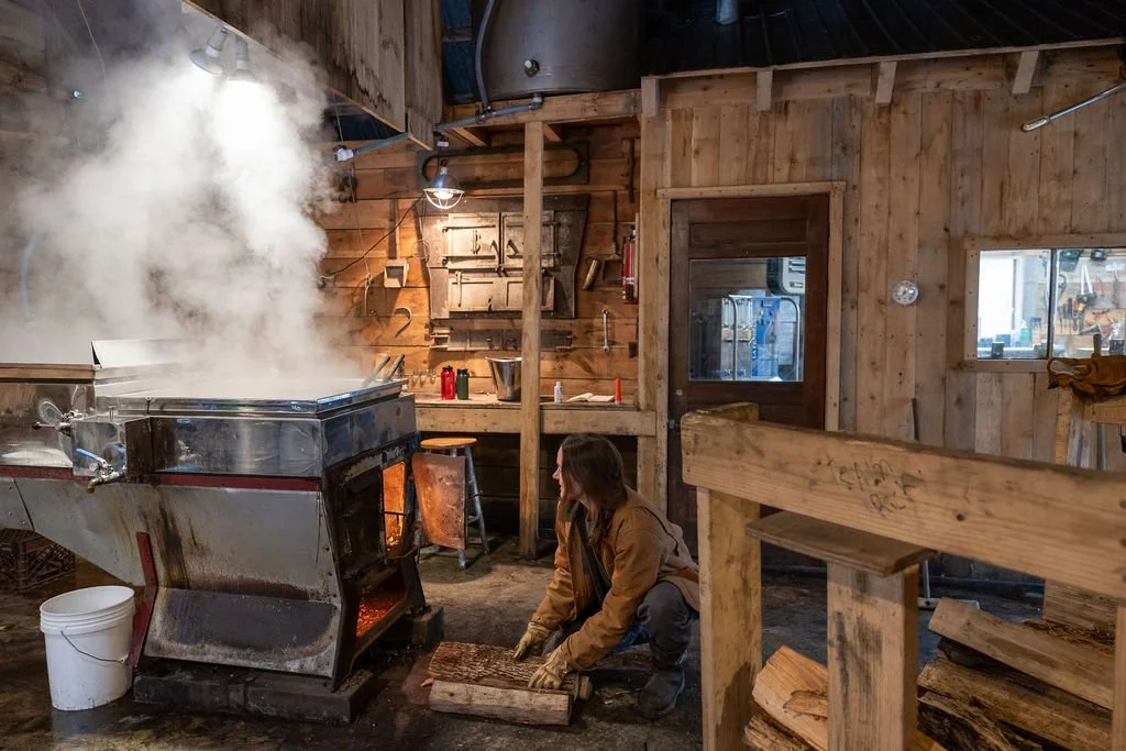 Person loading an arch with firewood inside a rustic wooden sugarhouse.