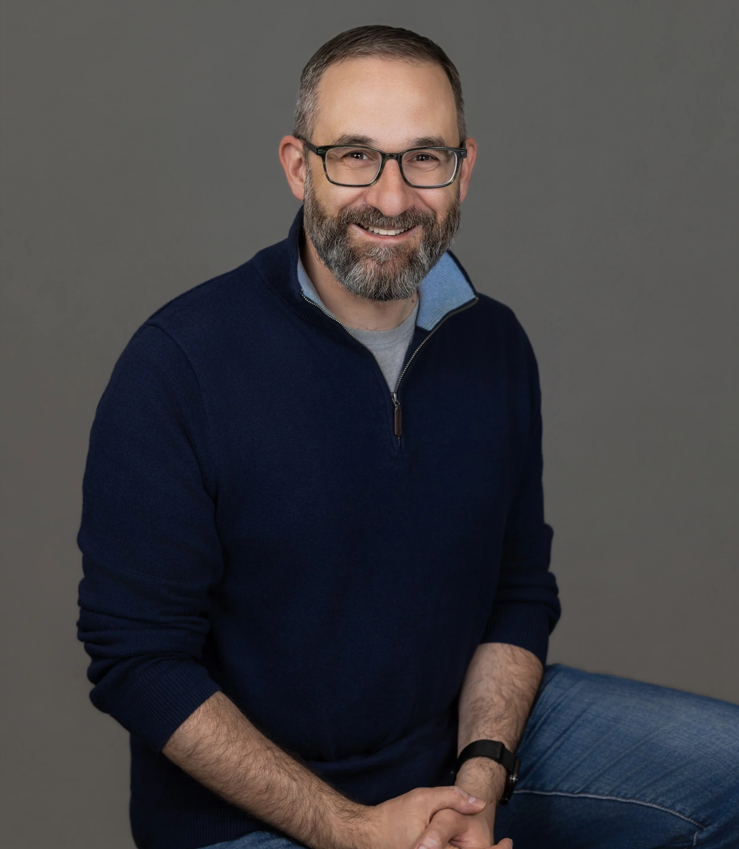 Smiling man with glasses and a beard wearing a navy blue sweater and gray shirt, posing against a gray background.