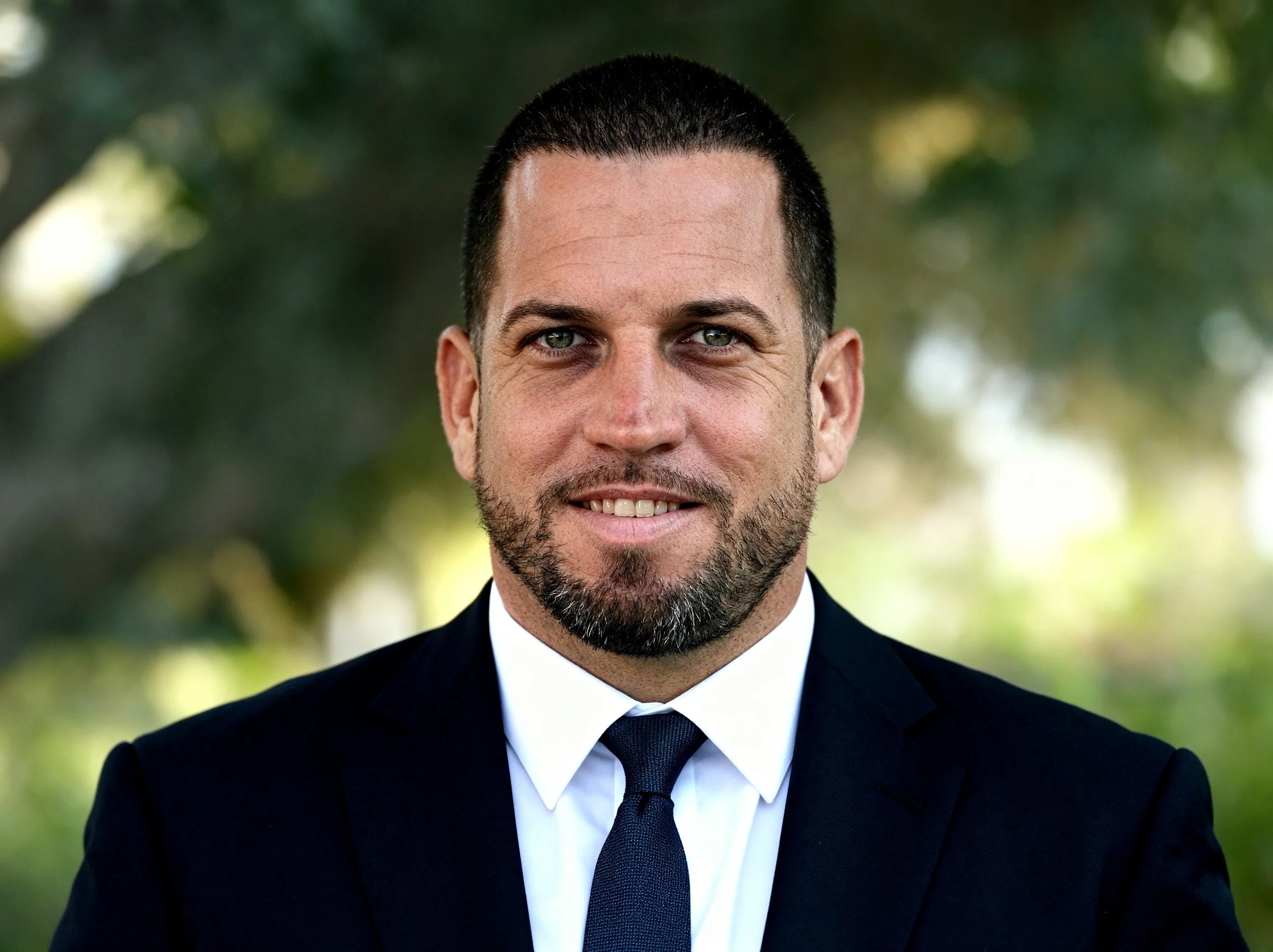 A man with short dark hair and a beard, wearing a dark suit, white shirt, and dark tie, smiling outdoors with blurred trees in the background.