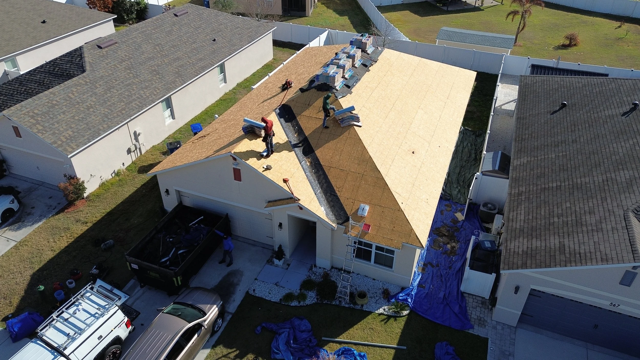 Aerial view of a house under construction, showing workers installing a new roof with plywood sheathing, surrounded by neighboring houses, parked vehicles, and construction materials.