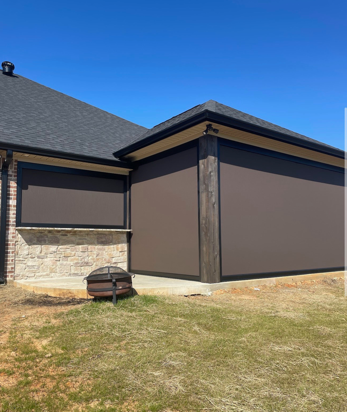 The image shows the exterior corner of a house with black siding, a stone foundation, and a gray roof. There is a concrete patio and a fire pit in the yard. The sky is clear and blue.
