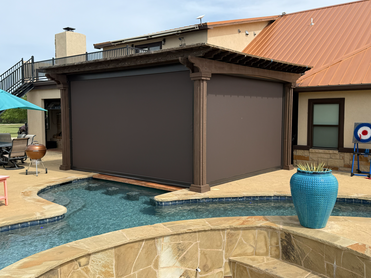 Backyard swimming pool with a closed brown pool cover, surrounded by beige stone patio, a blue planter with plants, outdoor furniture, and a house with beige walls and a copper-colored metal roof.