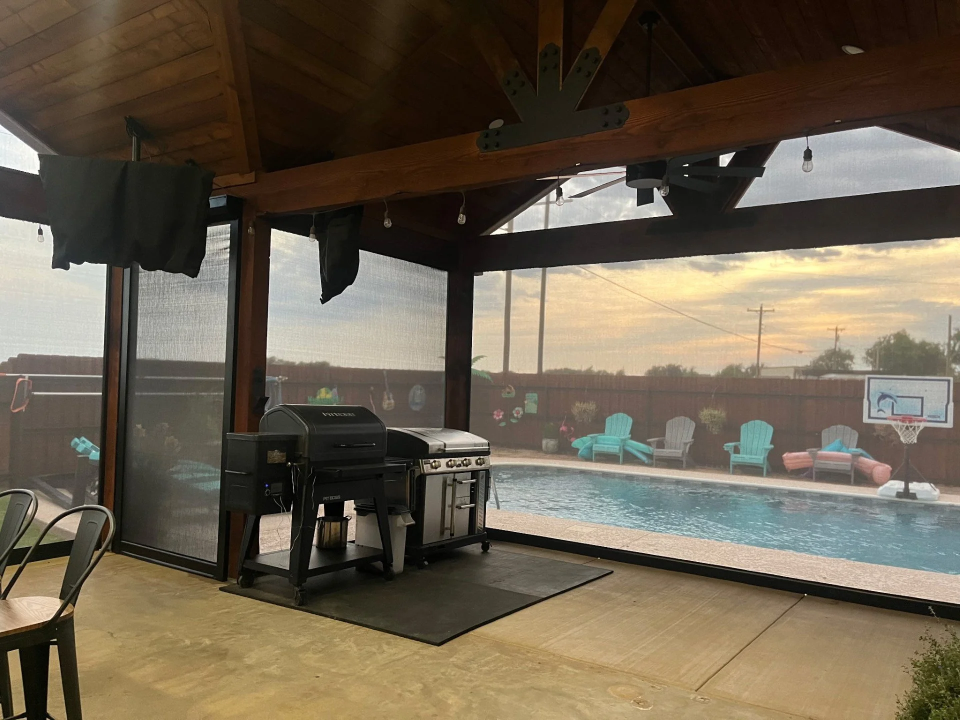 View of a backyard swimming pool area with outdoor chairs, a basketball hoop, and a fenced yard with trees, viewed through a screened porch with ceiling fans and string lights.
