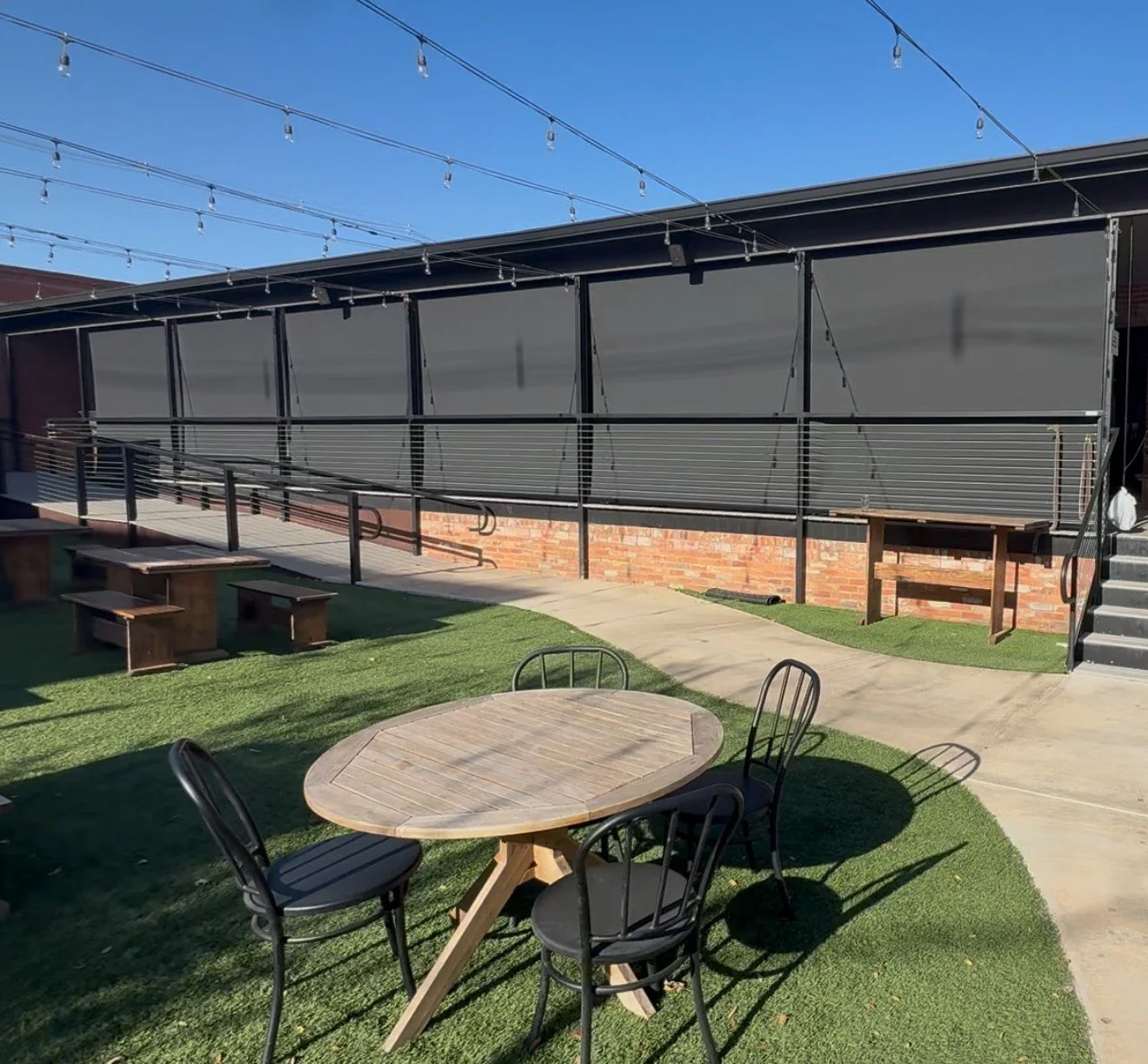 An outdoor patio area with a round wooden table and four black chairs on a grassy surface. There are string lights overhead, and a building with black exterior walls, metal railing, and stairs on the right side.
