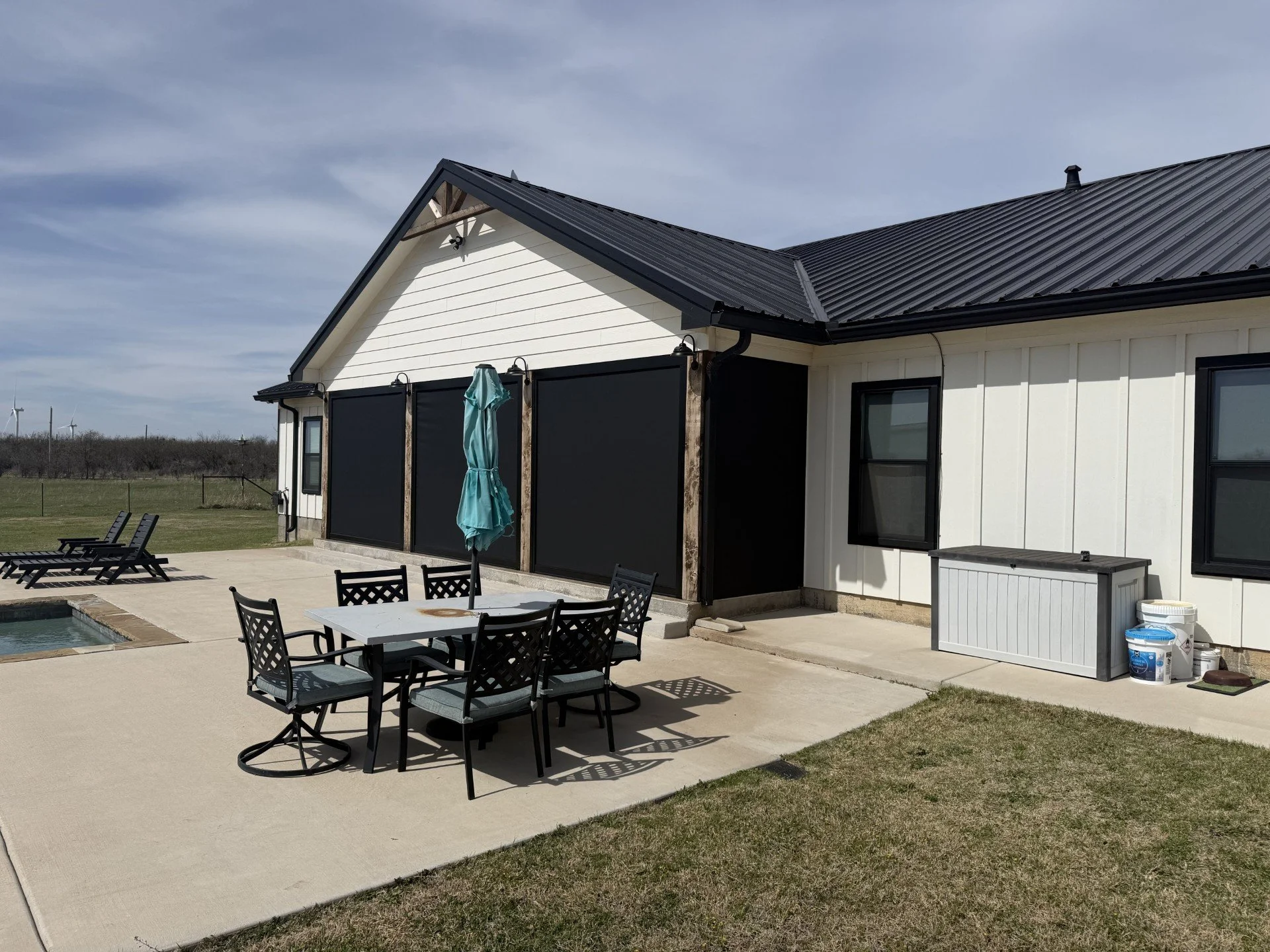 An outdoor patio area with black dining chairs and a white table under a patio umbrella next to a swimming pool. There is a white house with black trim and a black metal roof, with wind turbines in the background.