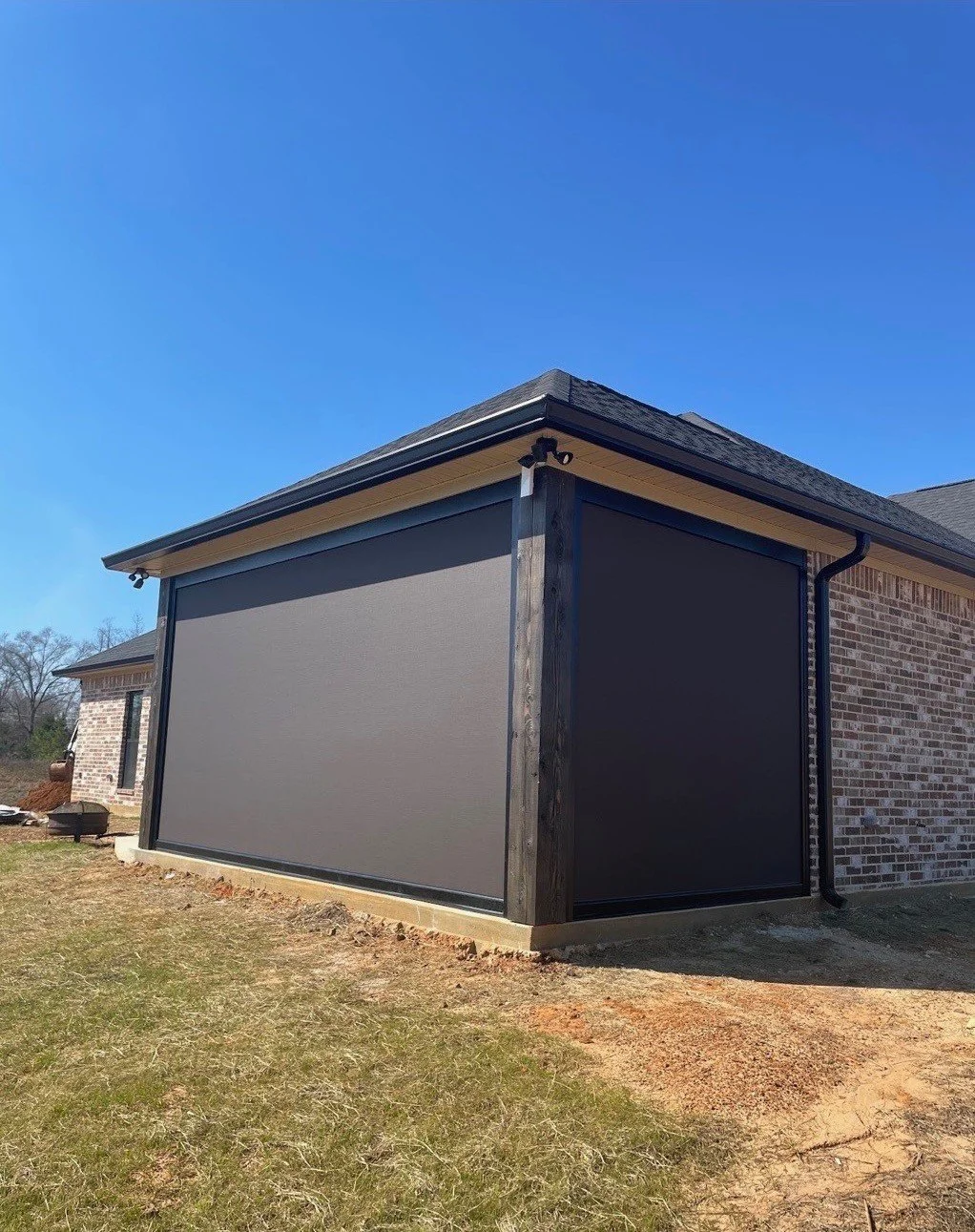 Side view of a house showing a newly installed retractable screen porch with dark gray fabric, black frame, and a brick exterior on a clear, sunny day.
