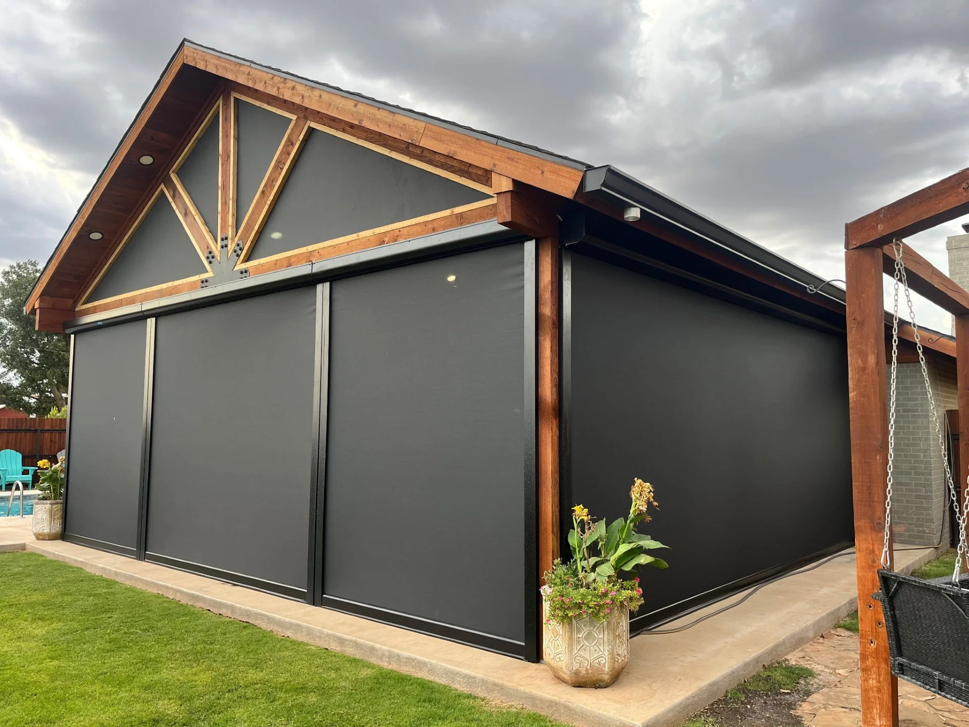 Backyard patio with a house featuring a black screen enclosure, a wooden pergola with redwood beams, potted plants, and outdoor furniture, under a cloudy sky.