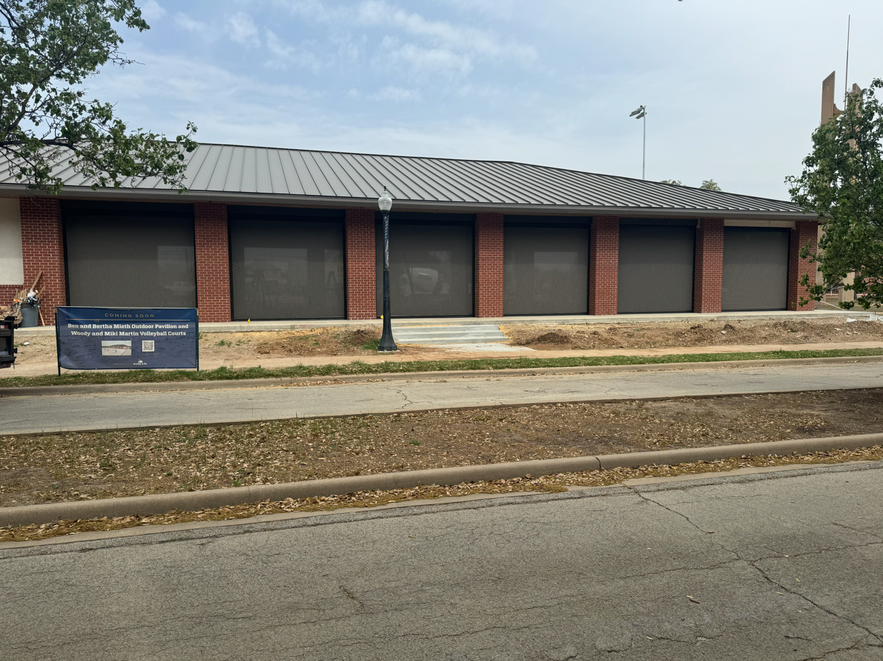 A brick building with five large closed black roller shutters on the front. There is a sign on the left side indicating the opening of a new outdoor pavilion and volleyball courts. A street runs in front with a sidewalk and a lamppost in the middle.