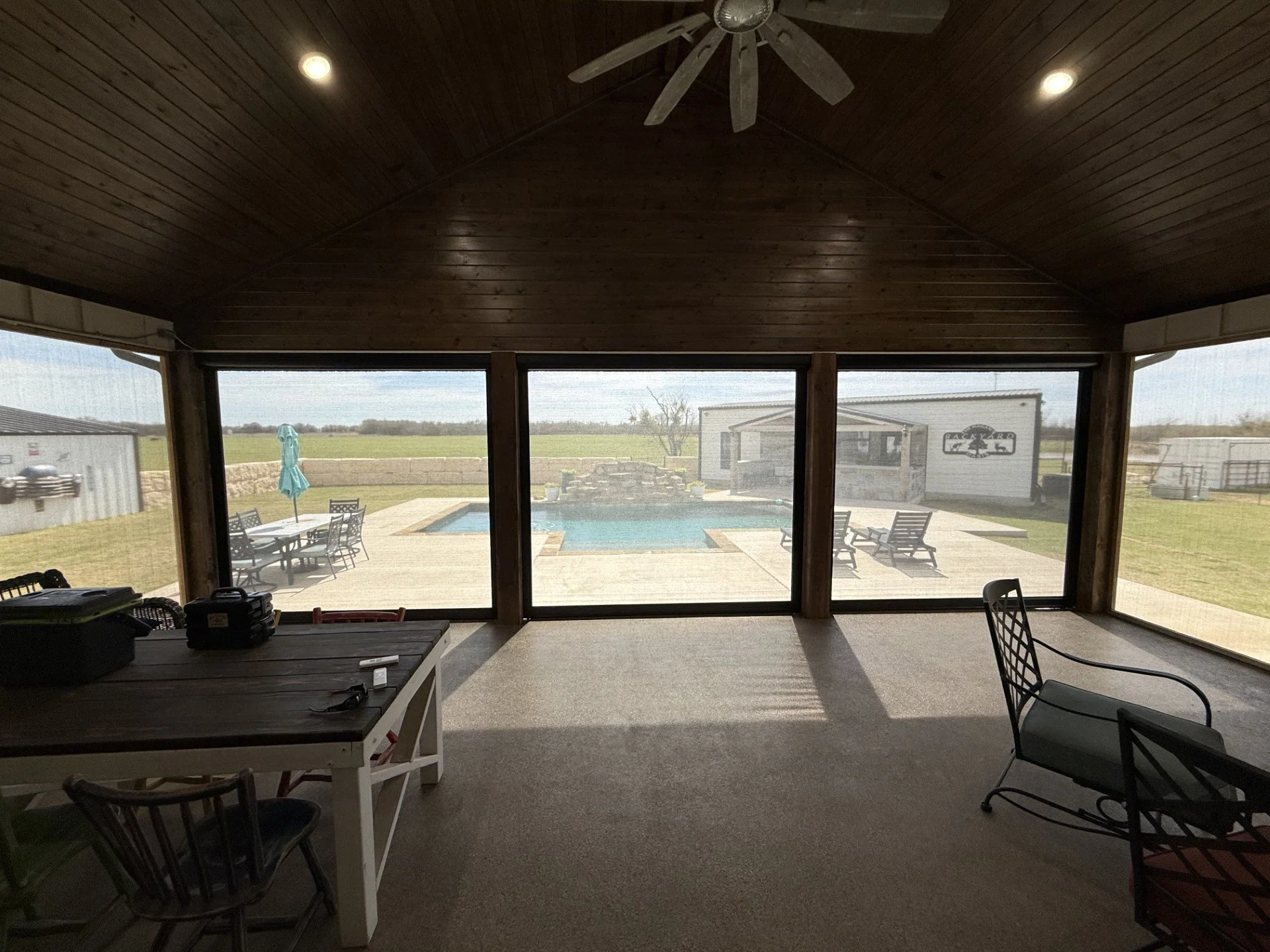 View from inside a screened porch looking out at a backyard with a swimming pool, patio furniture, a shed, and a grassy field beyond.