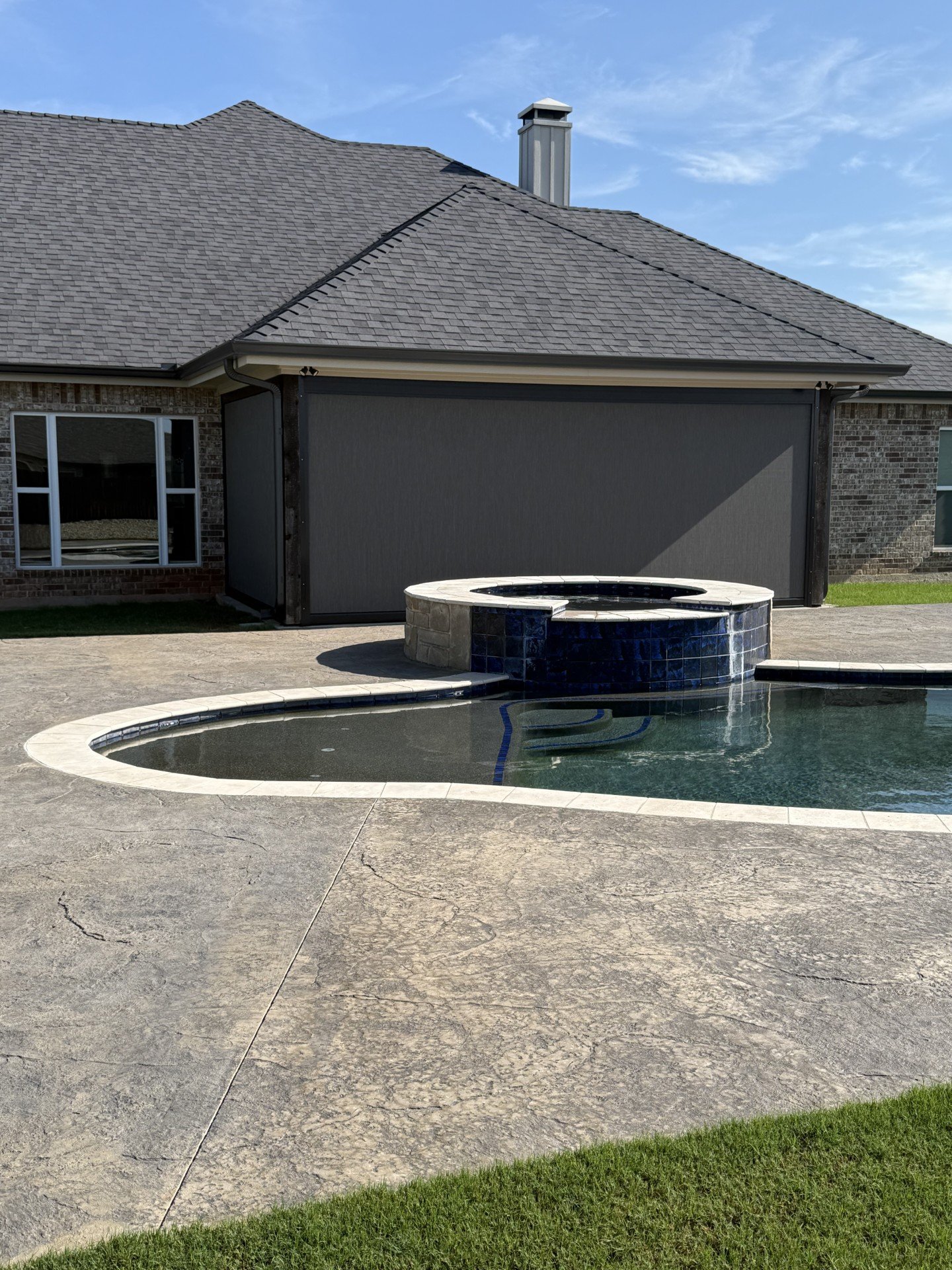 Backyard with a swimming pool with a built-in hot tub and a concrete deck, house with brick and gray siding, and a clear blue sky.