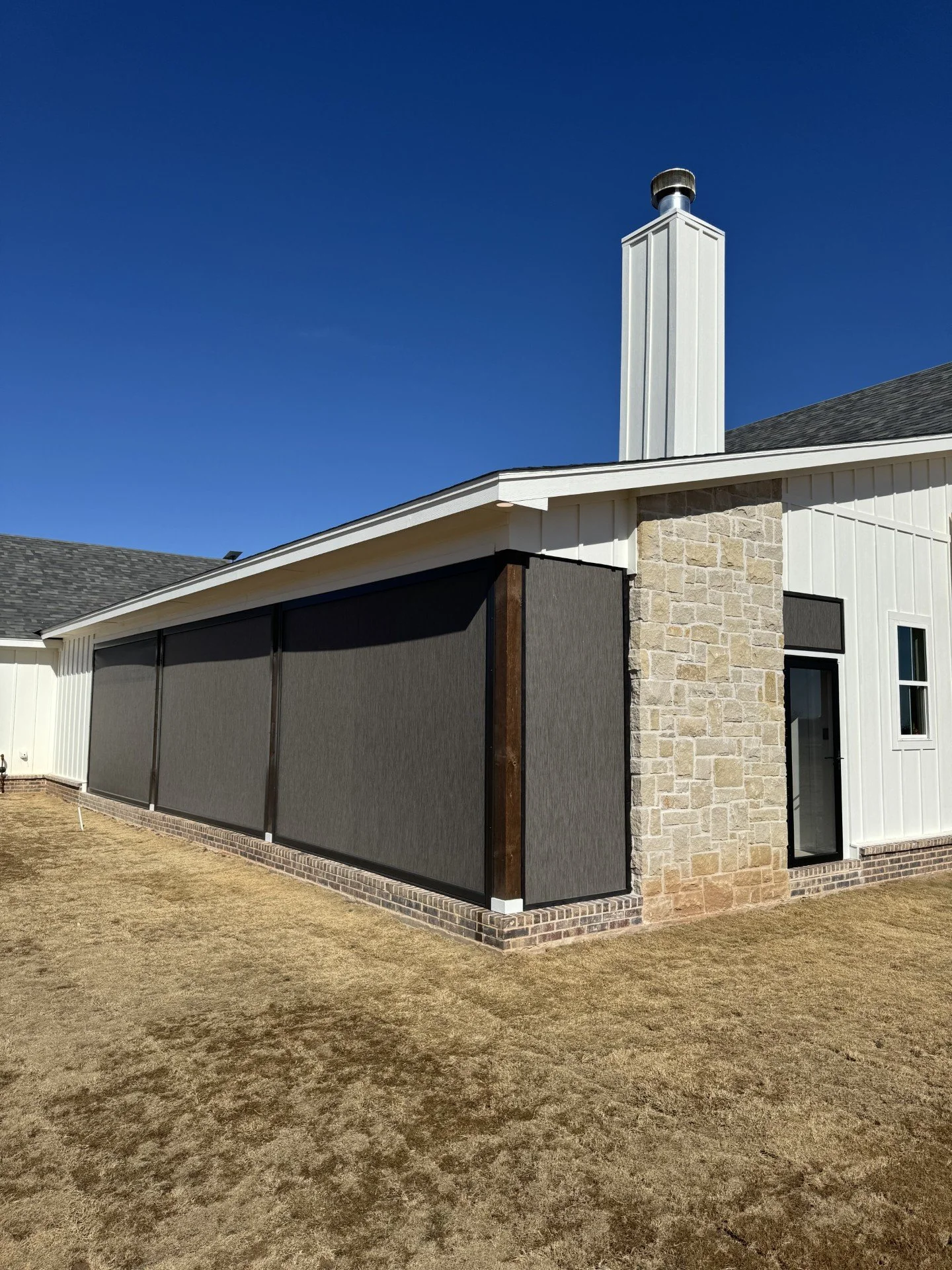 Backyard view of a house with a brick foundation, white siding, and a black window. There is a screened porch with dark mesh panels and a white roof trim. The ground is covered with dry grass, and the house has a stone chimney.