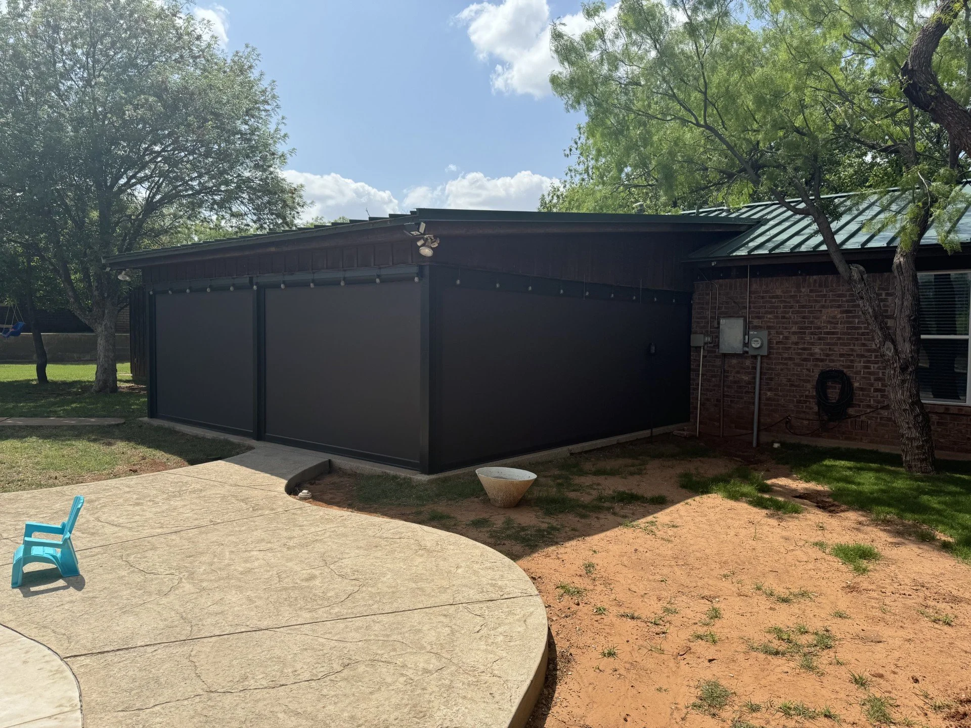 Backyard scene featuring a brick house with a green metal roof, trees, a patio with a small blue chair, and a black screened structure.