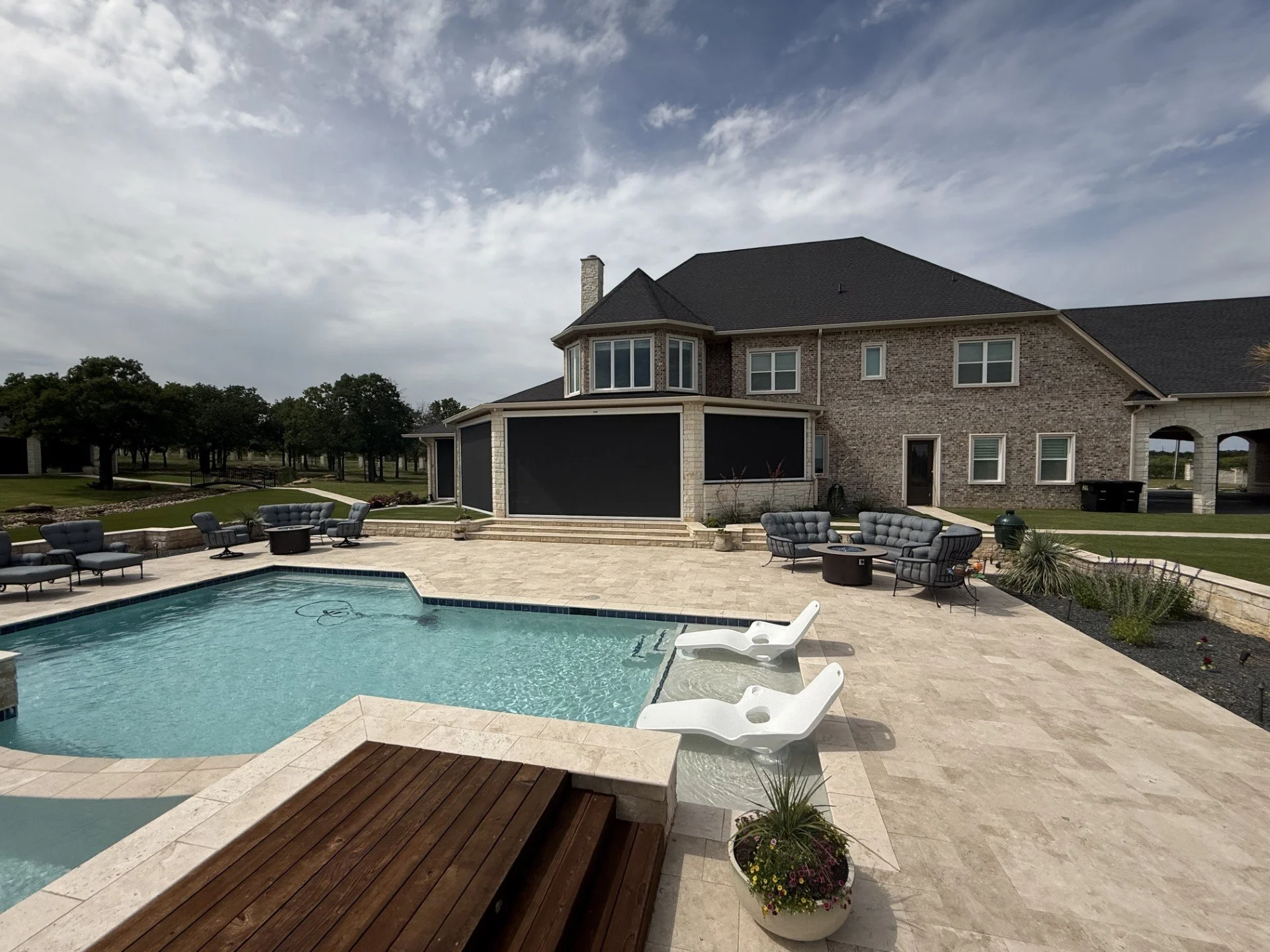Backyard of a house featuring a swimming pool surrounded by outdoor furniture, such as chairs, tables, and lounge chairs, with a brick house in the background, lush landscaping, and a partly cloudy sky overhead.