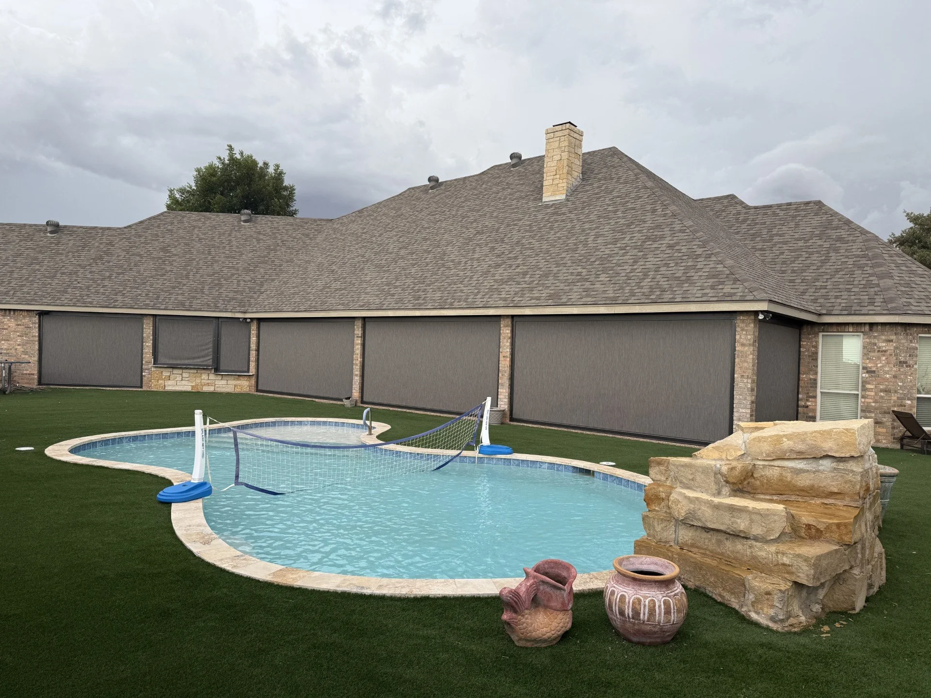 Backyard with swimming pool, artificial grass, large rocks, decorative vases, and a house with a peaked roof and brick exterior walls.