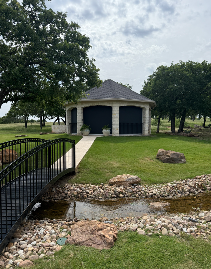 A small stone building with a dark roof and dark shutters, located in a landscaped park with a small creek, rocks, a bridge, and green trees.