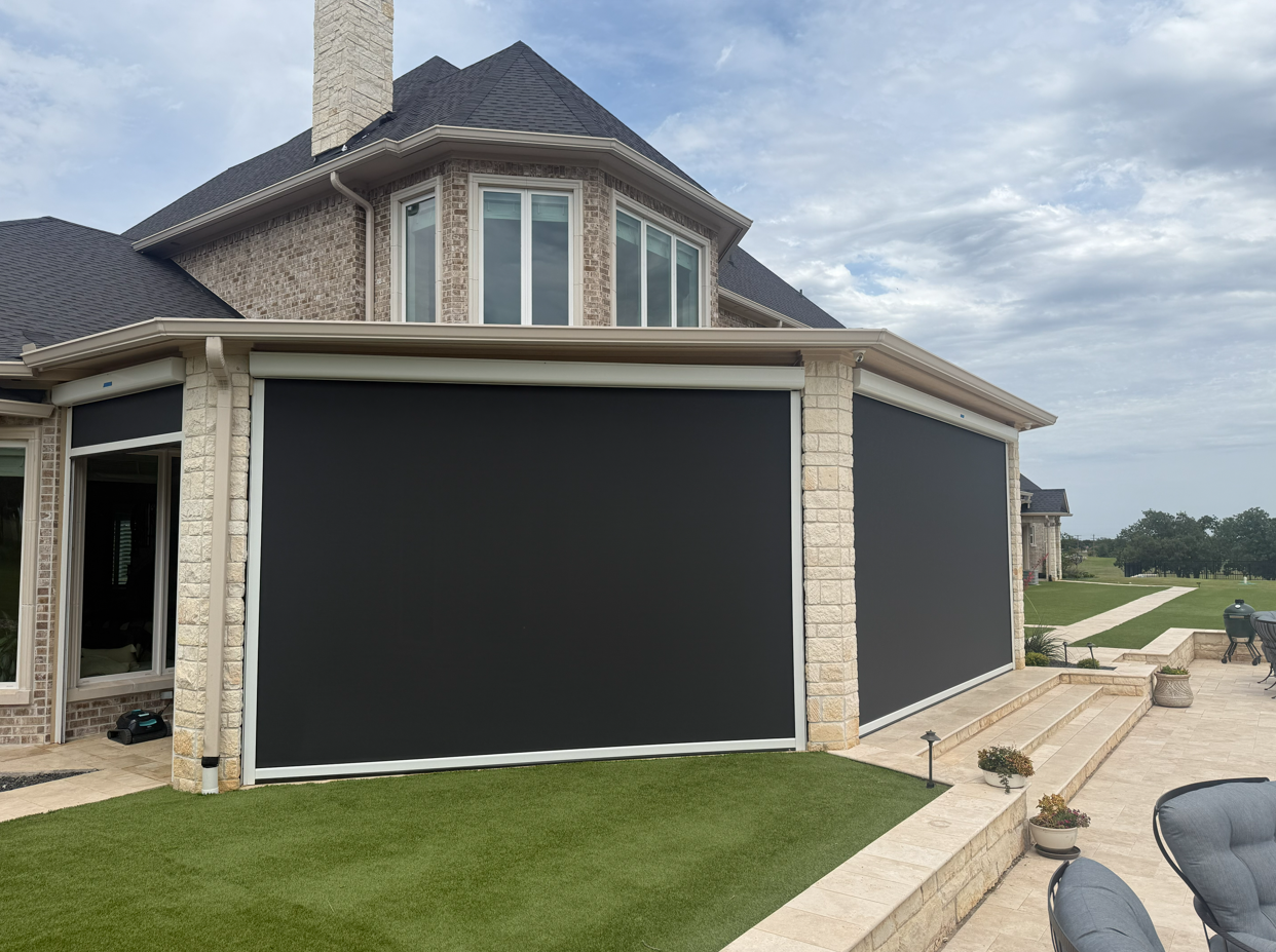 A large brick house with a patio and rolling lawn, featuring two black retractable screens or shades on the back porch.