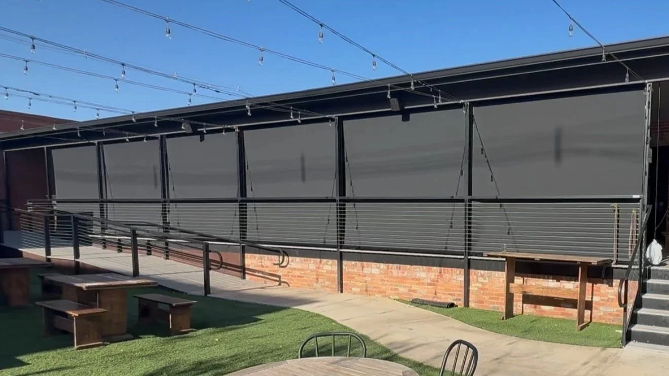 Outdoor patio area with folding black walls, string lights overhead, a ramp with black railings, brick wall, and wooden benches on grass and concrete surfaces.