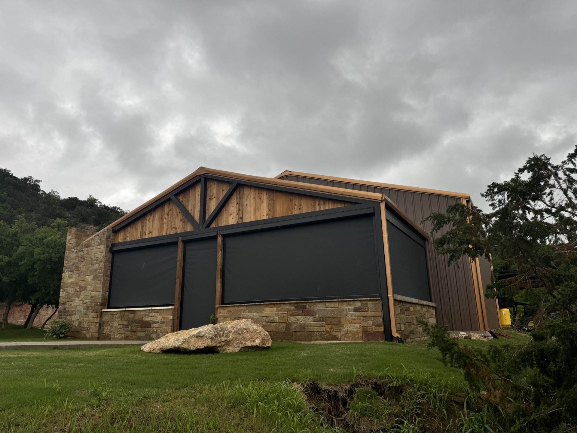 Modern building with a brick lower section and wooden upper section, located in a green landscape with trees and a large rock in the foreground, under a cloudy sky.