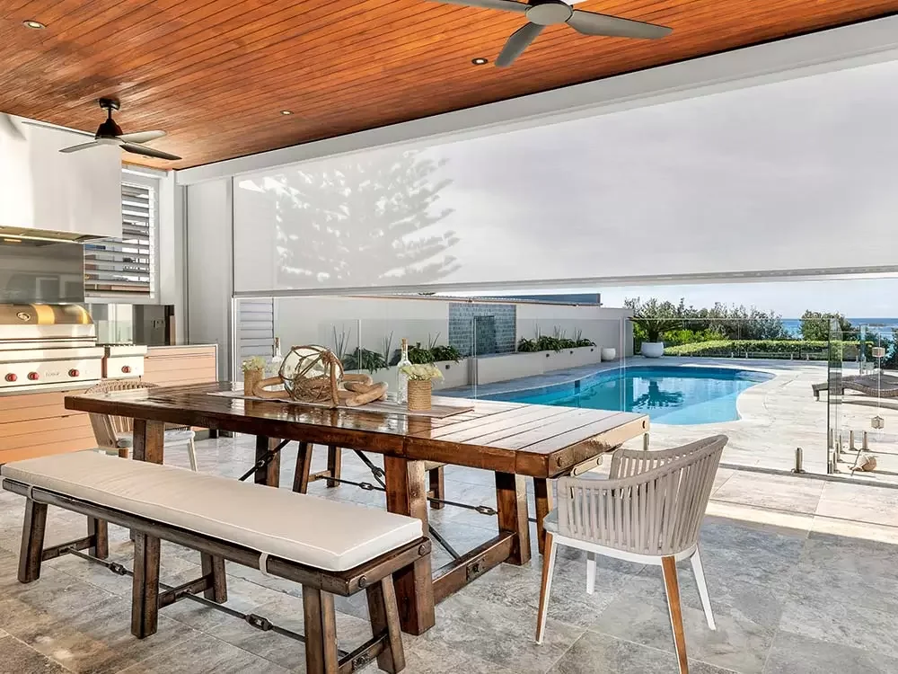 Indoor-outdoor living space with wooden ceiling, ceiling fans, dining table, bench, chair, and a pool with a view of the ocean.