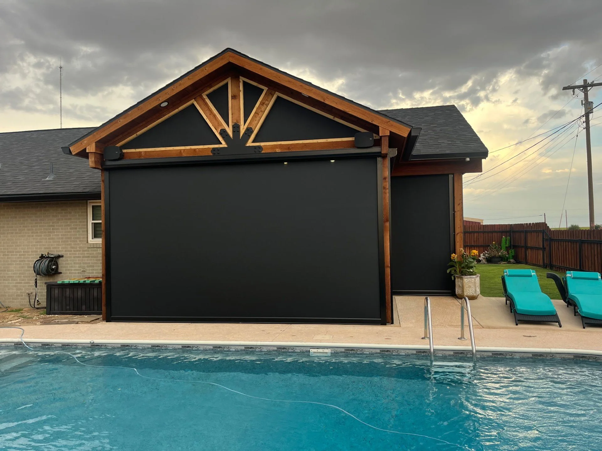 Backyard with swimming pool, black outdoor movie screen, poolside chairs, potted plants, and a house with a gabled roof under cloudy sky.