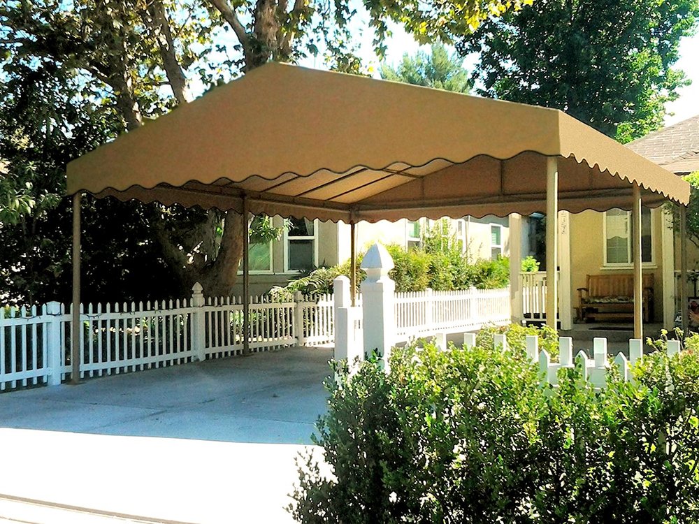 A beige canopy tent is set up over a concrete patio area in front of a house with a light yellow exterior, white picket fence, and green bushes. There is a wooden bench under the house's porch and trees in the background.