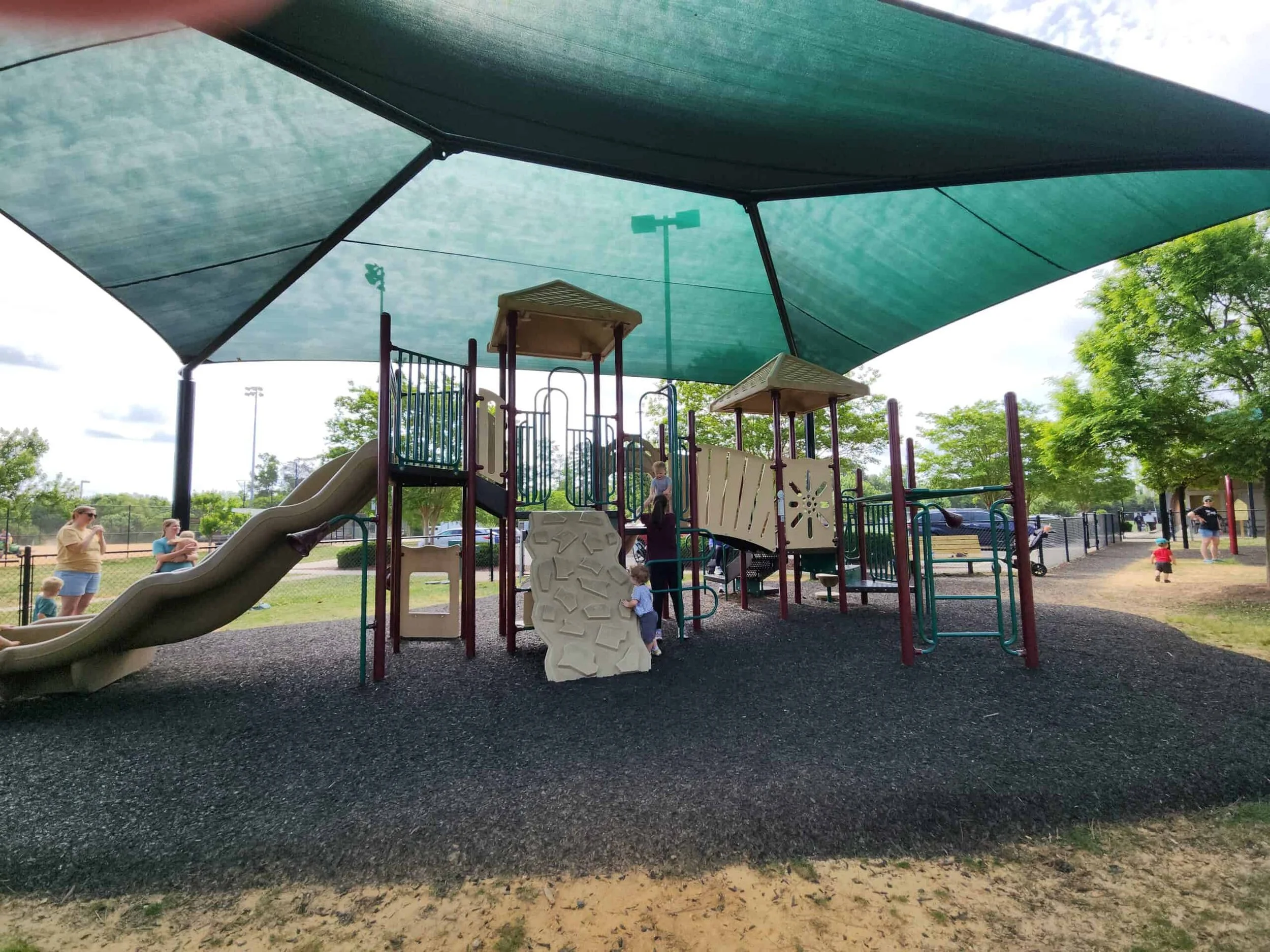 Children and adults are at a playground with a large shade canopy overhead. The playground has slides and climbing structures, with trees and a park setting in the background.