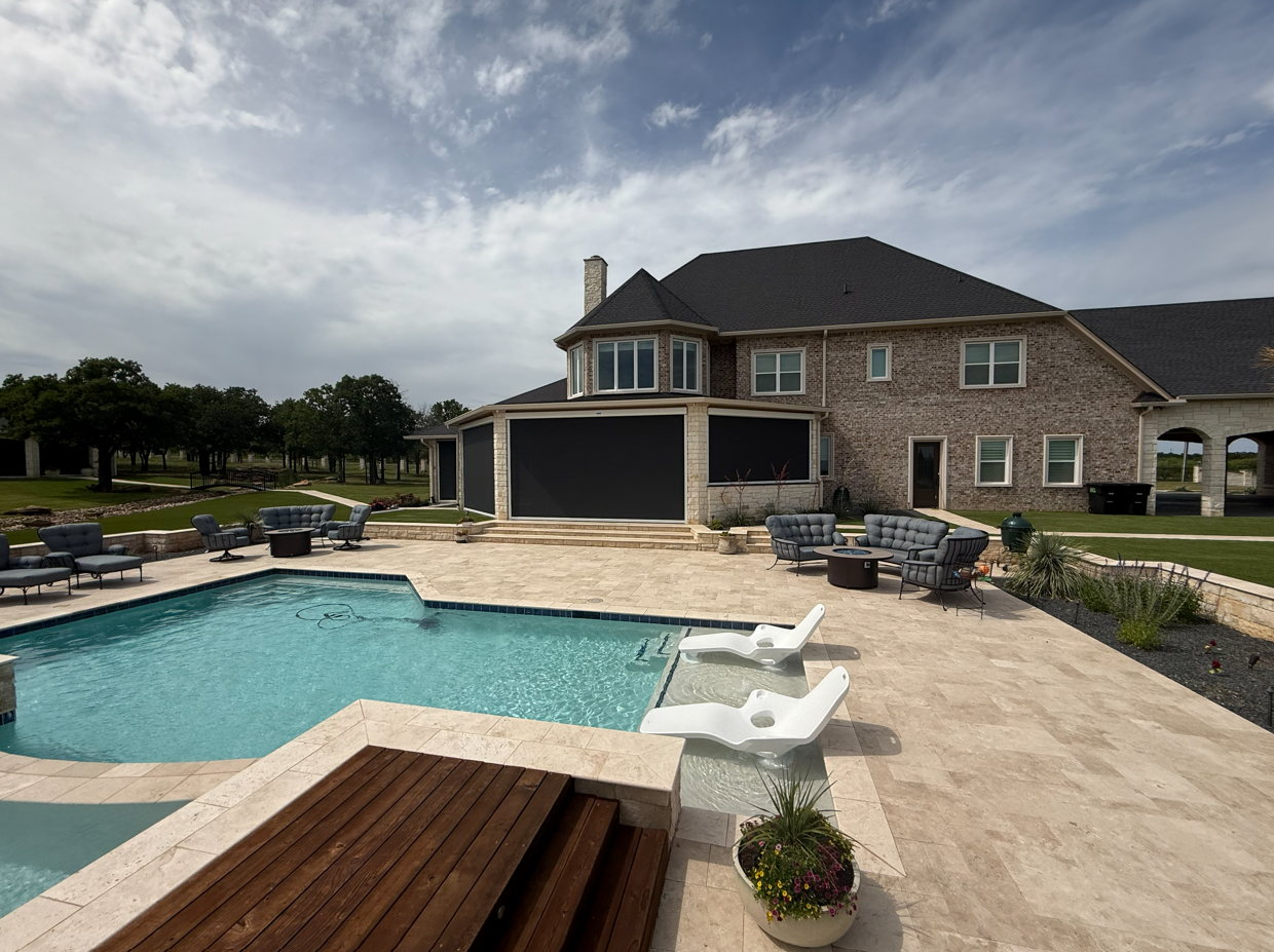 Backyard with a pool, patio furniture, and a large brick house under a partly cloudy sky.