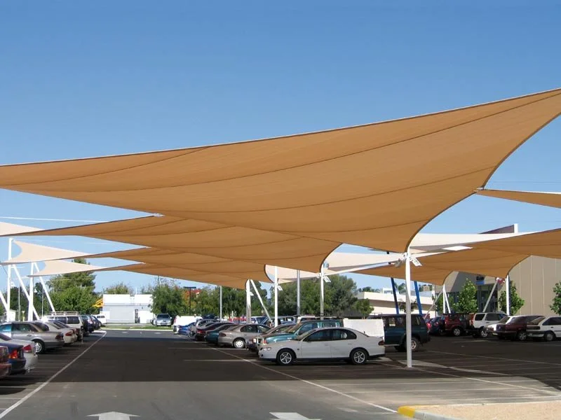 Parking lot with beige fabric shade sails overhead and cars parked underneath on a sunny day.