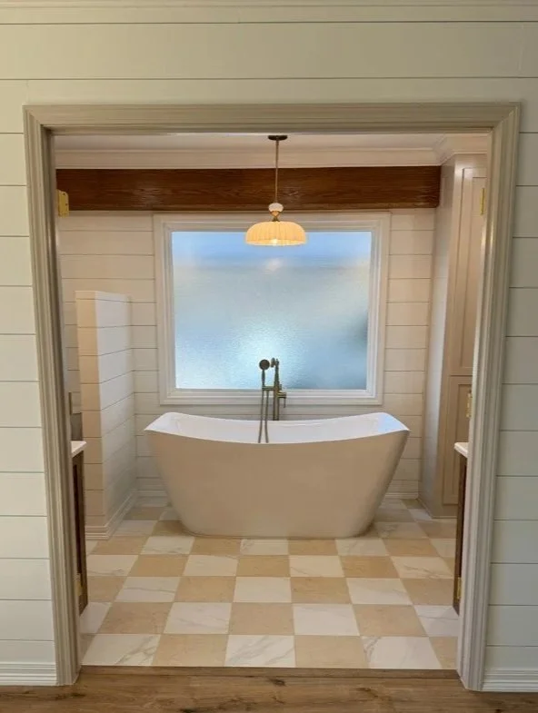 A freestanding bathtub in a bathroom, placed in front of a frosted window, with a pendant light hanging from the ceiling and wooden beams above.