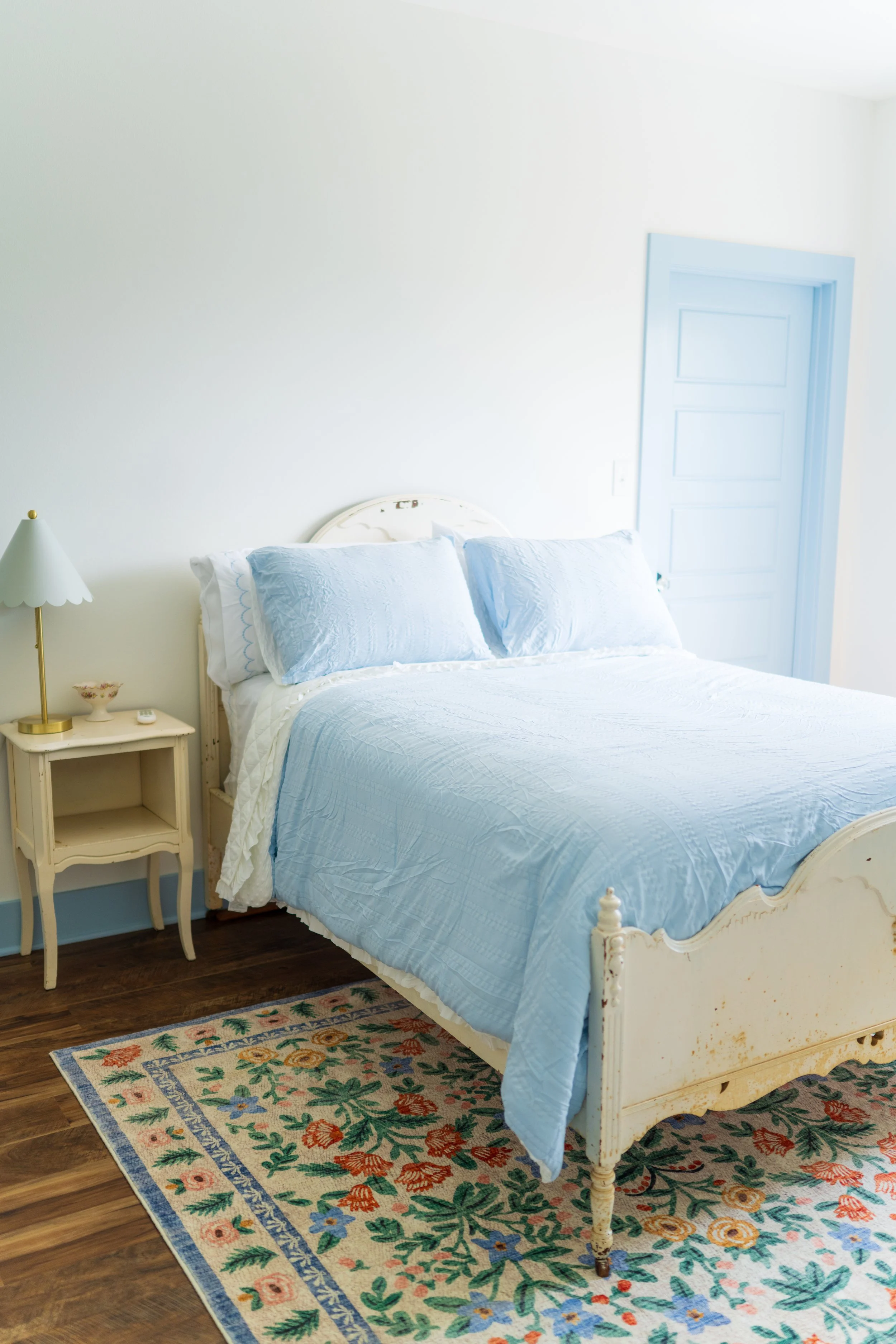 A cozy bedroom with a white vintage-style bed, light blue quilt, matching blue pillows, a beige nightstand with a lamp and small decorative bowl, floral area rug, wooden floors, and a light blue door.