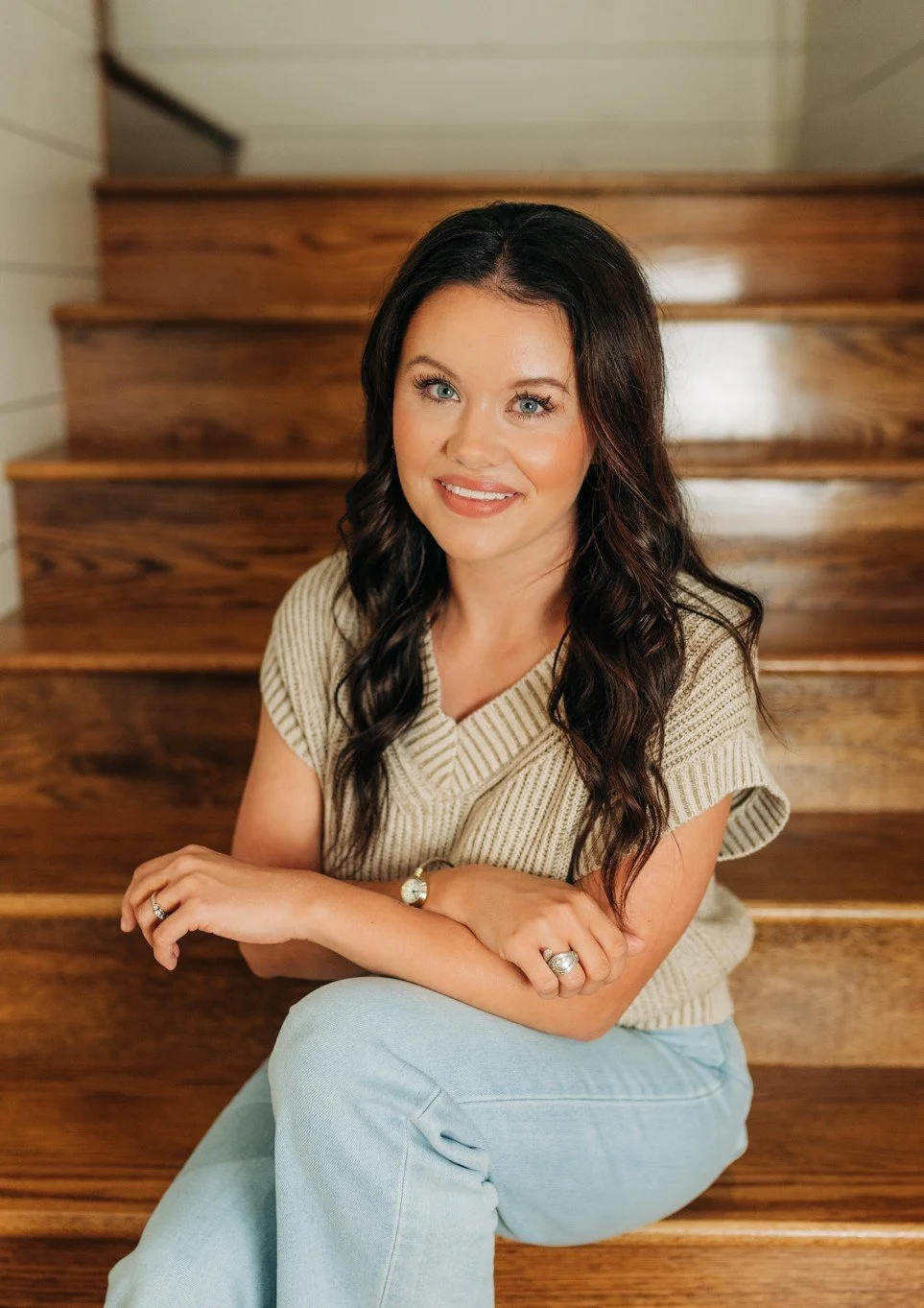A woman with long dark wavy hair and fair skin sitting on wooden stairs, smiling, wearing a beige short-sleeve knit top and light blue jeans, with jewelry including rings and a watch.