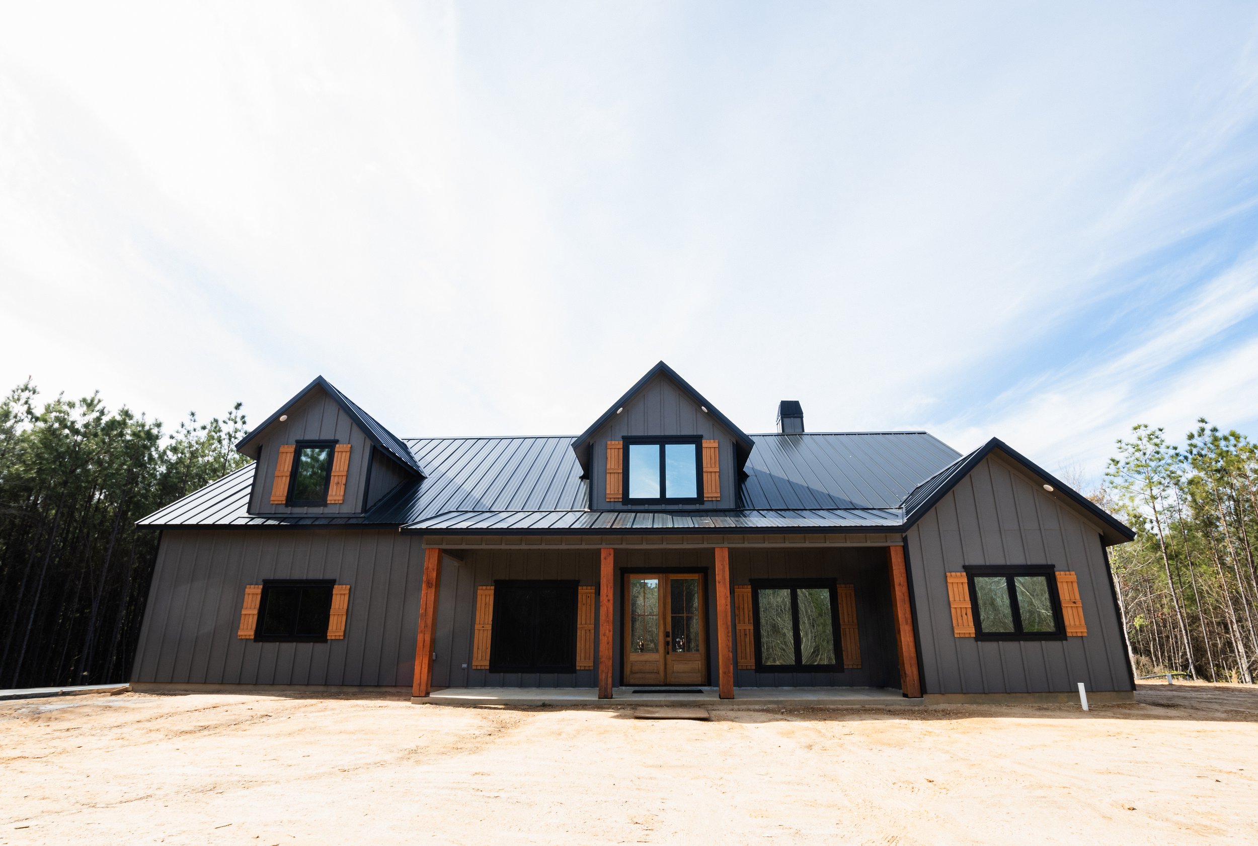 Front view of a modern house with black metal roof, gray siding, and wooden accents, set against a partly cloudy sky and surrounded by trees.