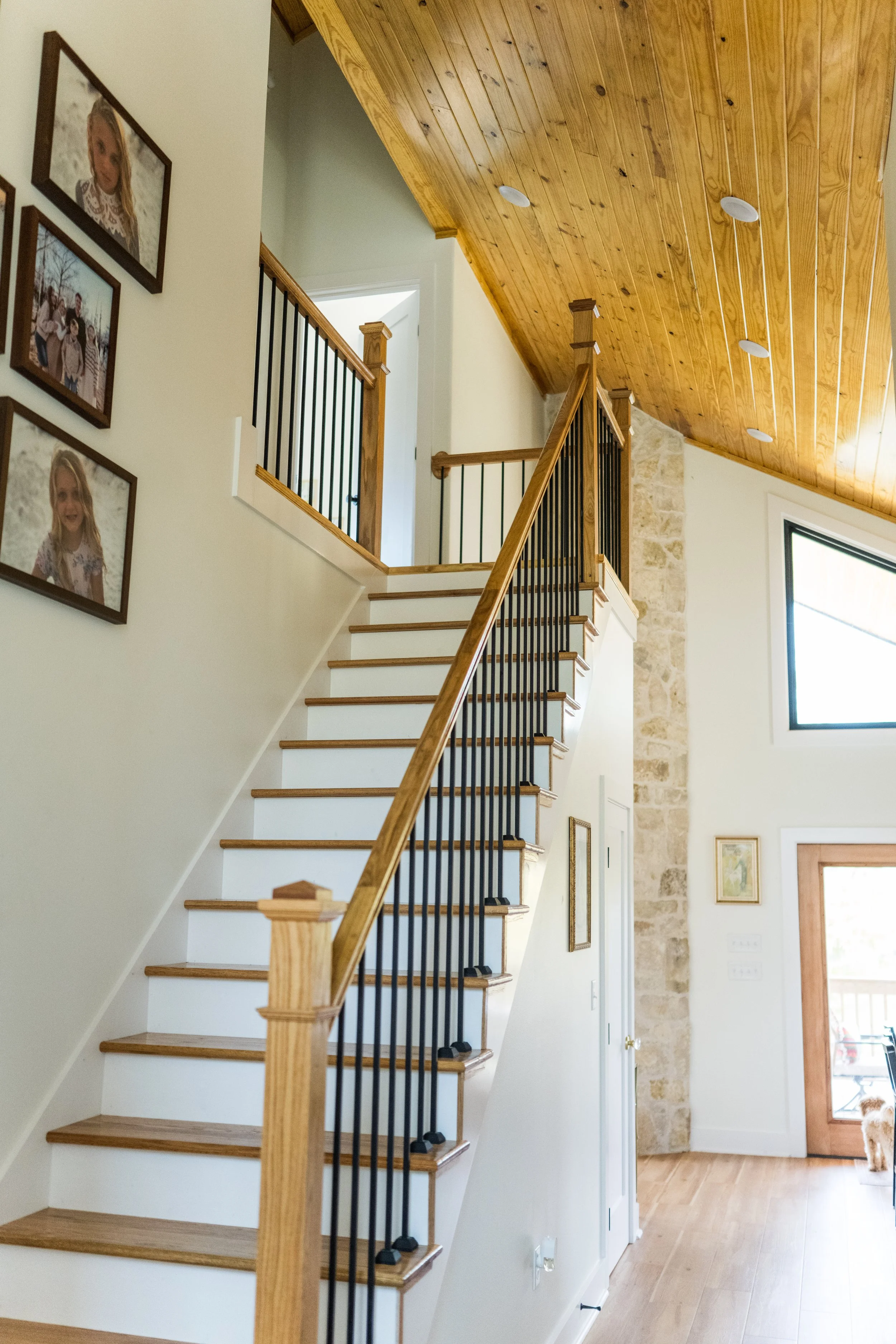 Interior view of a staircase in a house with wooden steps and black metal railings, leading to an upper level. The wall beside the staircase features framed photos of children. The ceiling is made of wood paneling with recessed lighting. At the bottom right, a dog is partially visible near a door and window.