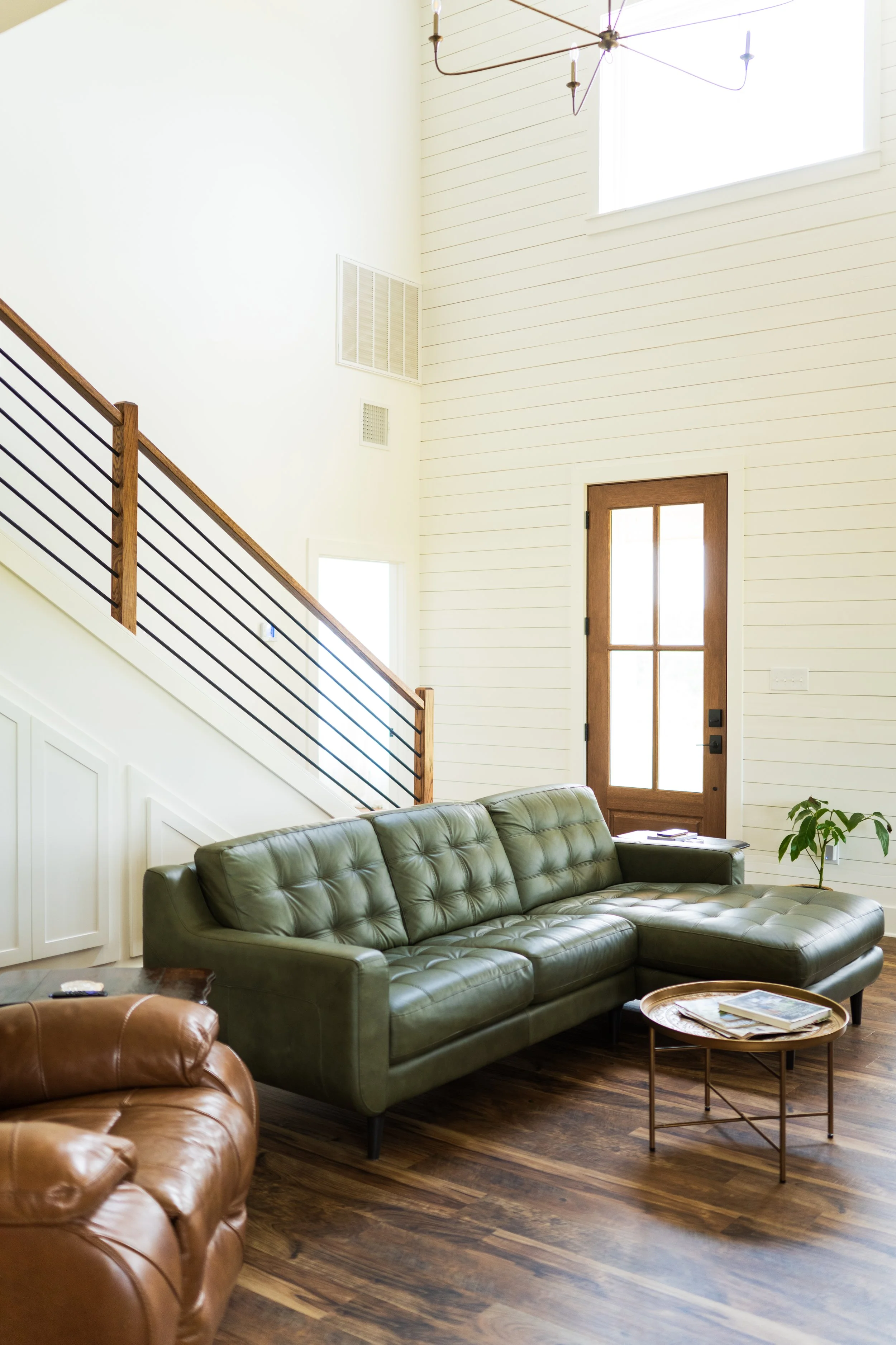 Living room with a green leather sofa, wooden floor, wooden door with glass panels, staircase with wooden handrail, and a white wall with high window letting in natural light.
