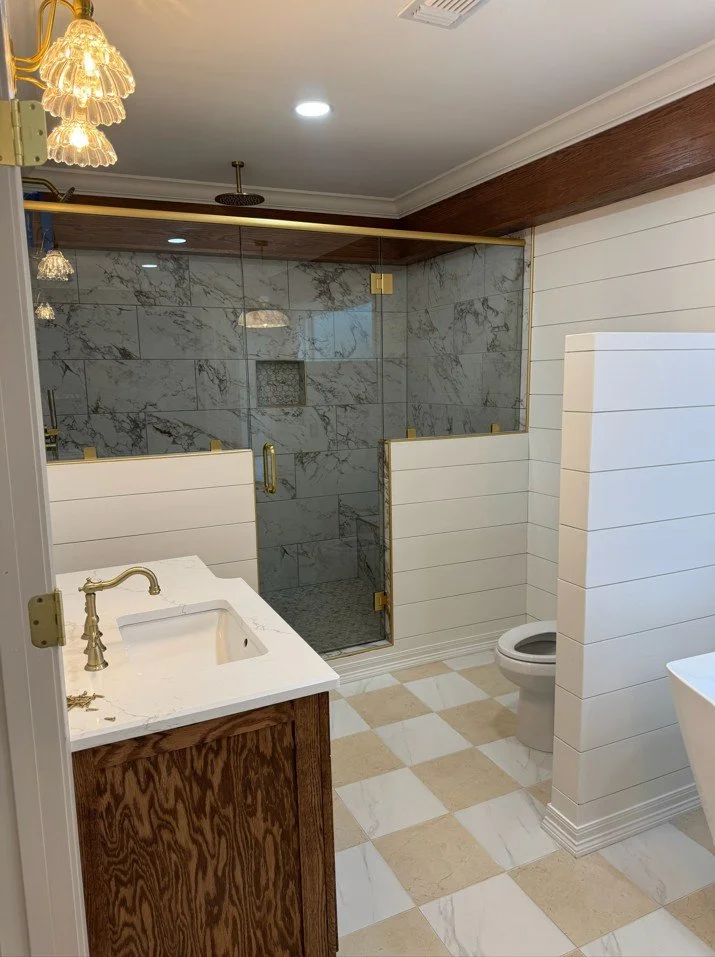 Modern bathroom with marble shower, white paneled walls, wooden accents, gold fixtures, and a beige and white checkered floor.