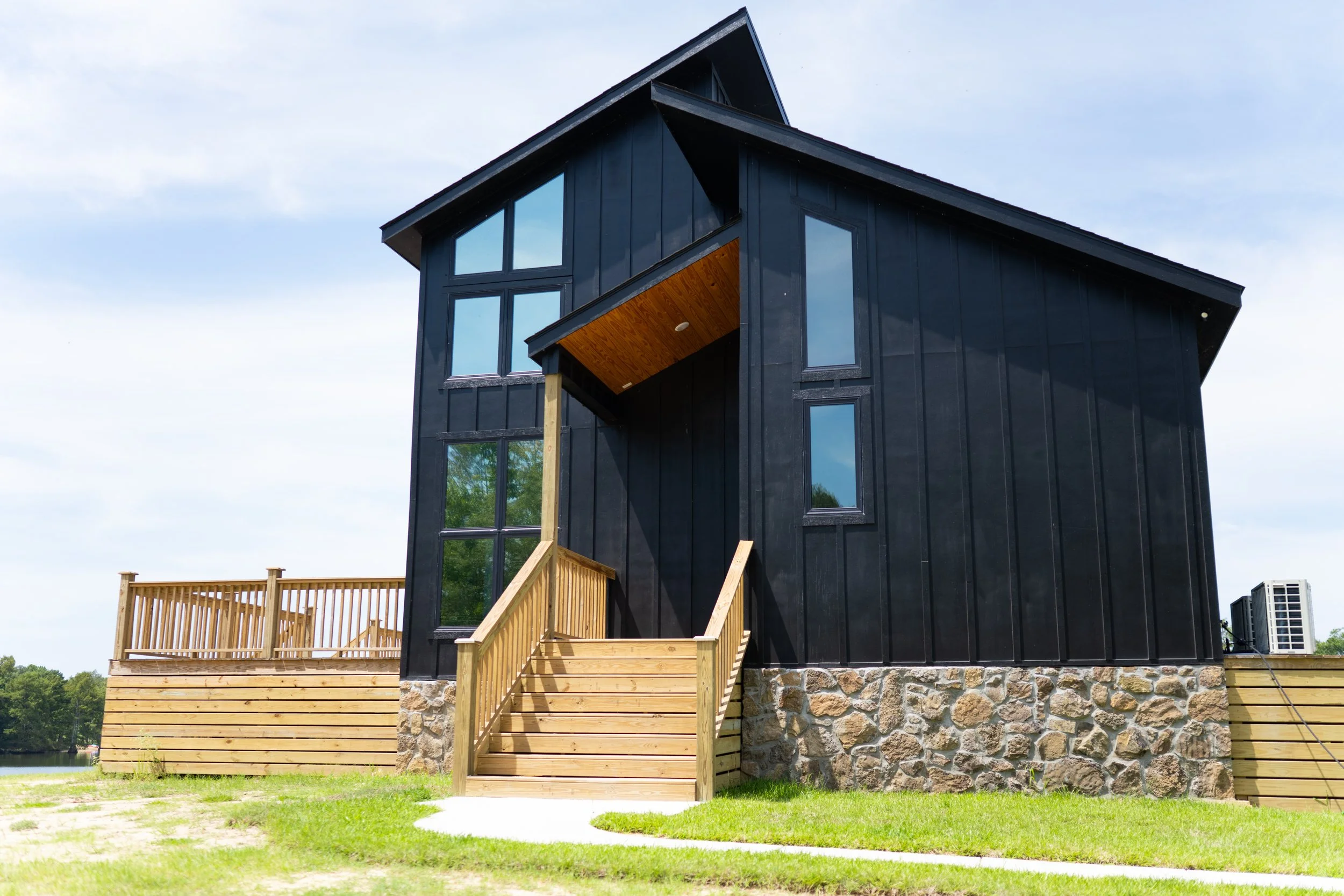 Black modern house with large windows, wooden porch, and stone foundation near a body of water.
