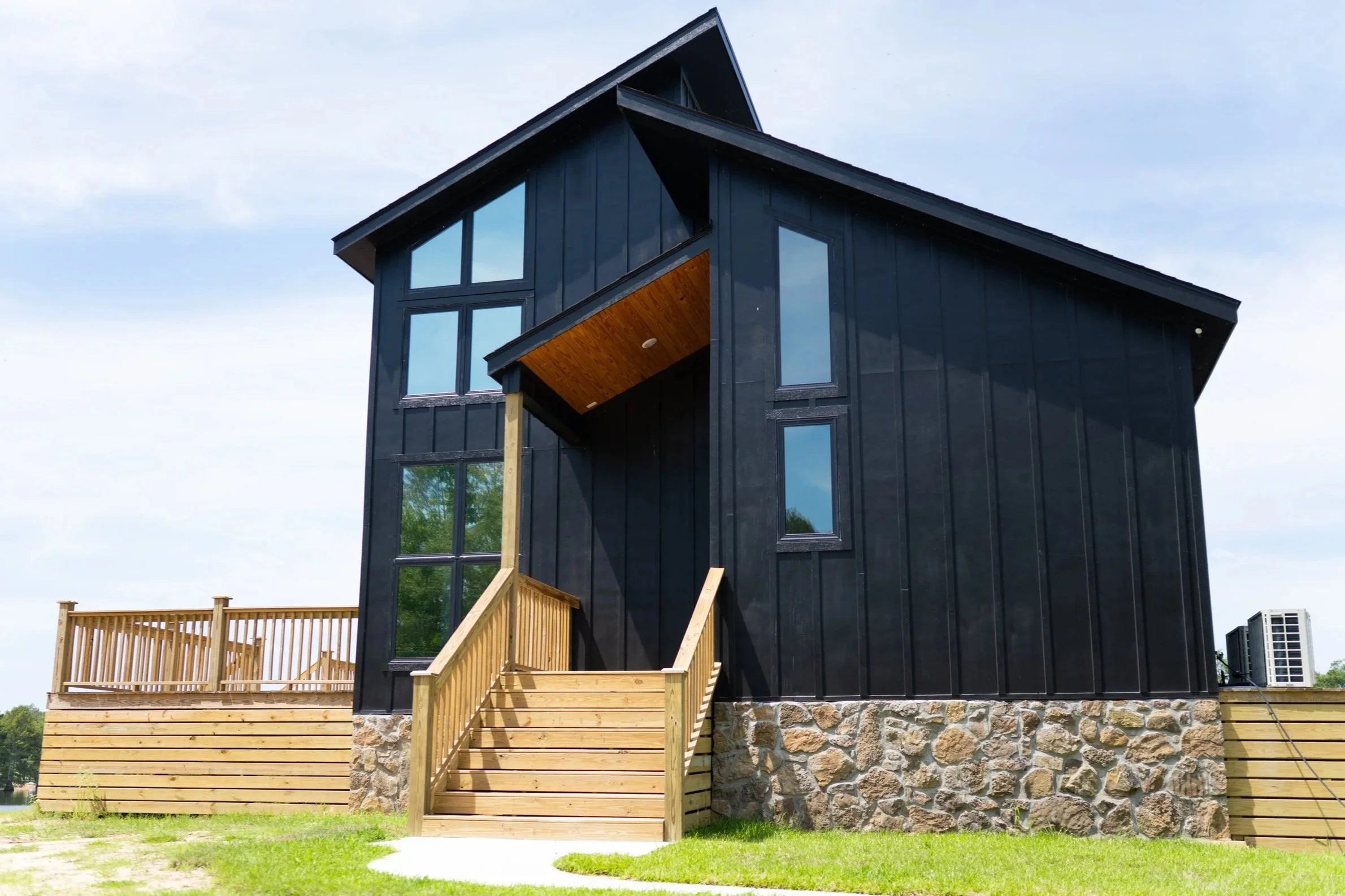 Modern black house with large windows and wooden deck under construction, with steps leading up to the deck and a stone foundation.