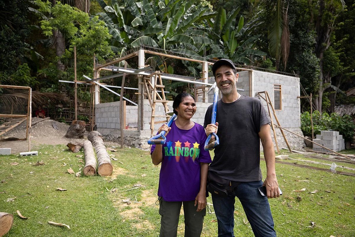 Two smiling people holding construction tools in front of a partially built house surrounded by lush green trees.