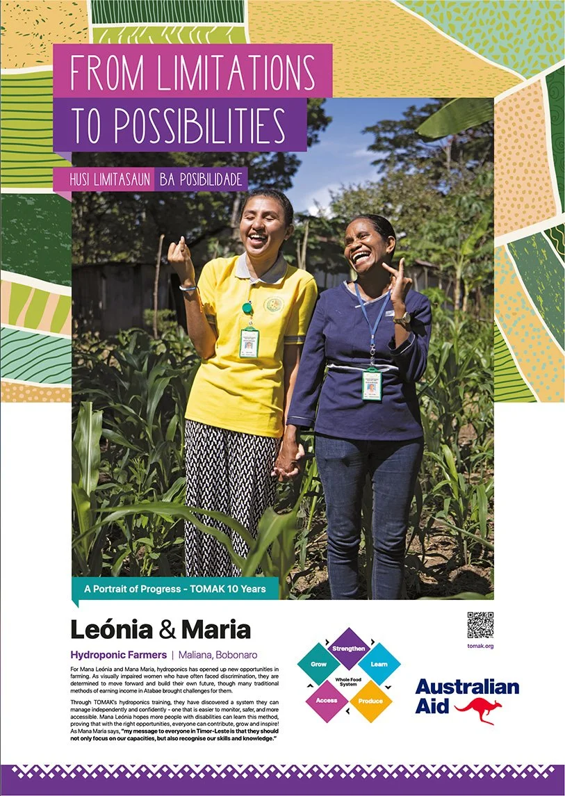 Two women standing outdoors in a farm, smiling and holding hands. One woman is wearing a yellow shirt, and the other is wearing a dark blue shirt. They are surrounded by green plants and trees, with a bright blue sky in the background.