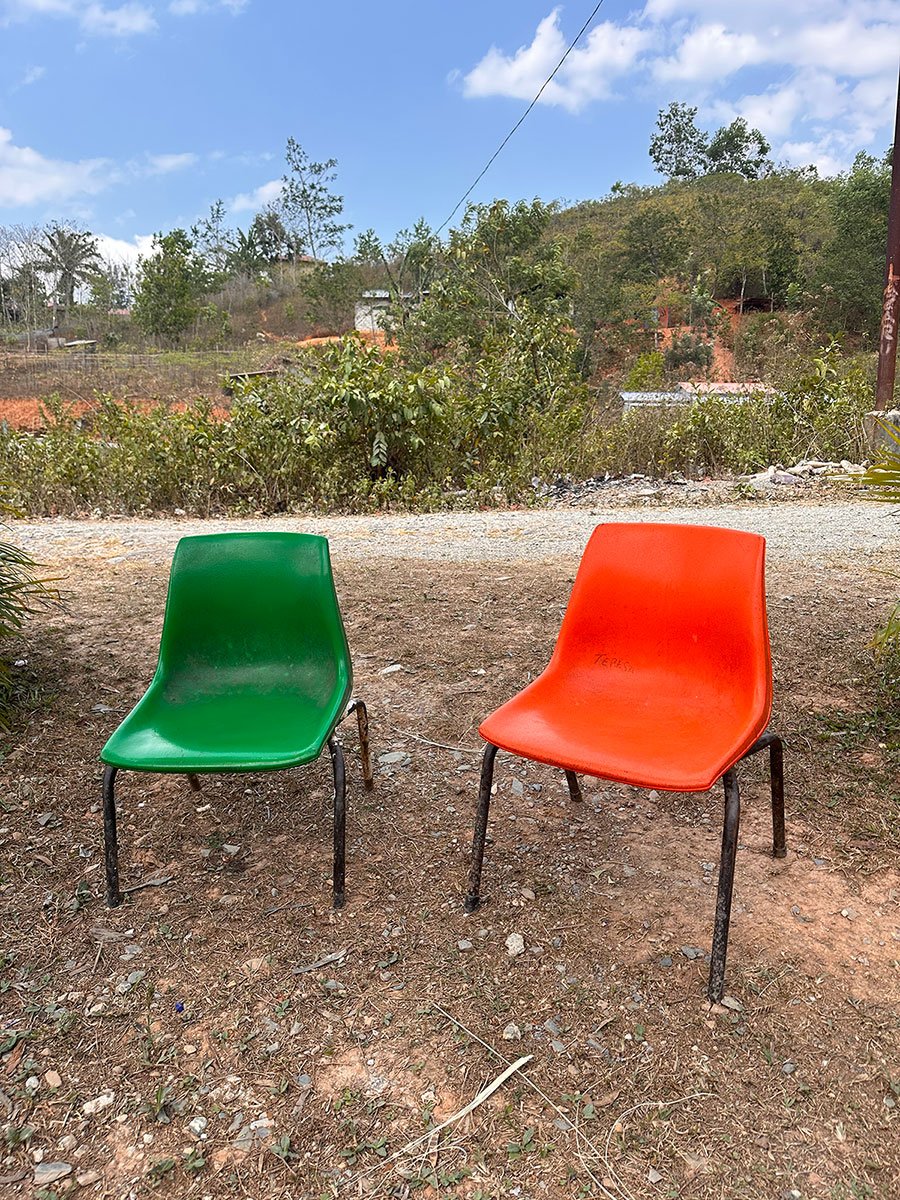 Two plastic chairs, one green and one red, placed on dirt ground outdoors with greenery and hills in the background.